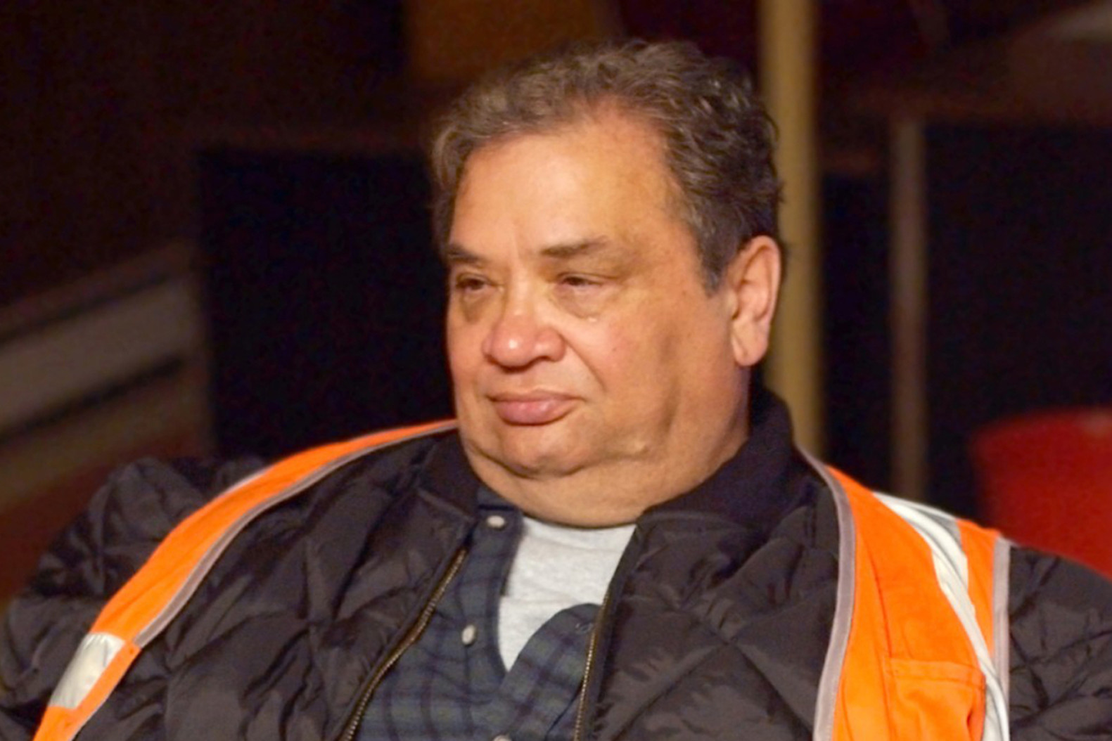 A man wearing an orange safety vest and a dark jacket, sitting indoors with a neutral expression.
