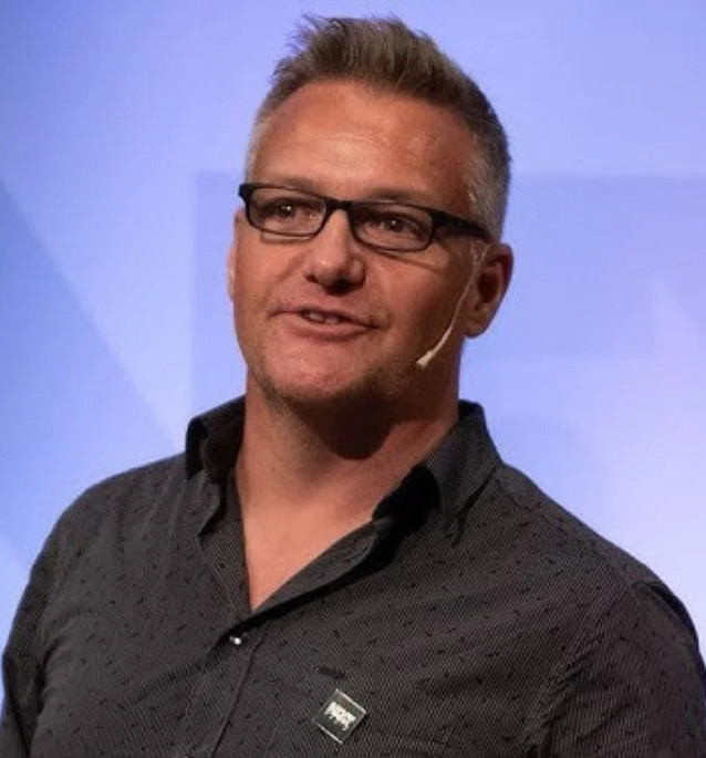 A man with glasses and a microphone, wearing a dark shirt, speaking on stage.