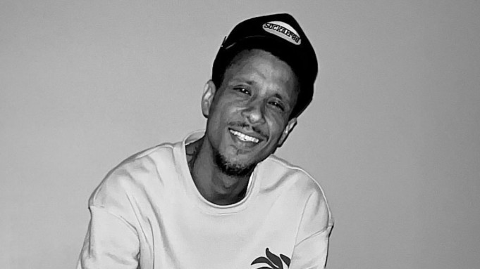 A man smiling, wearing a cap and a sweatshirt, in a black and white photo.