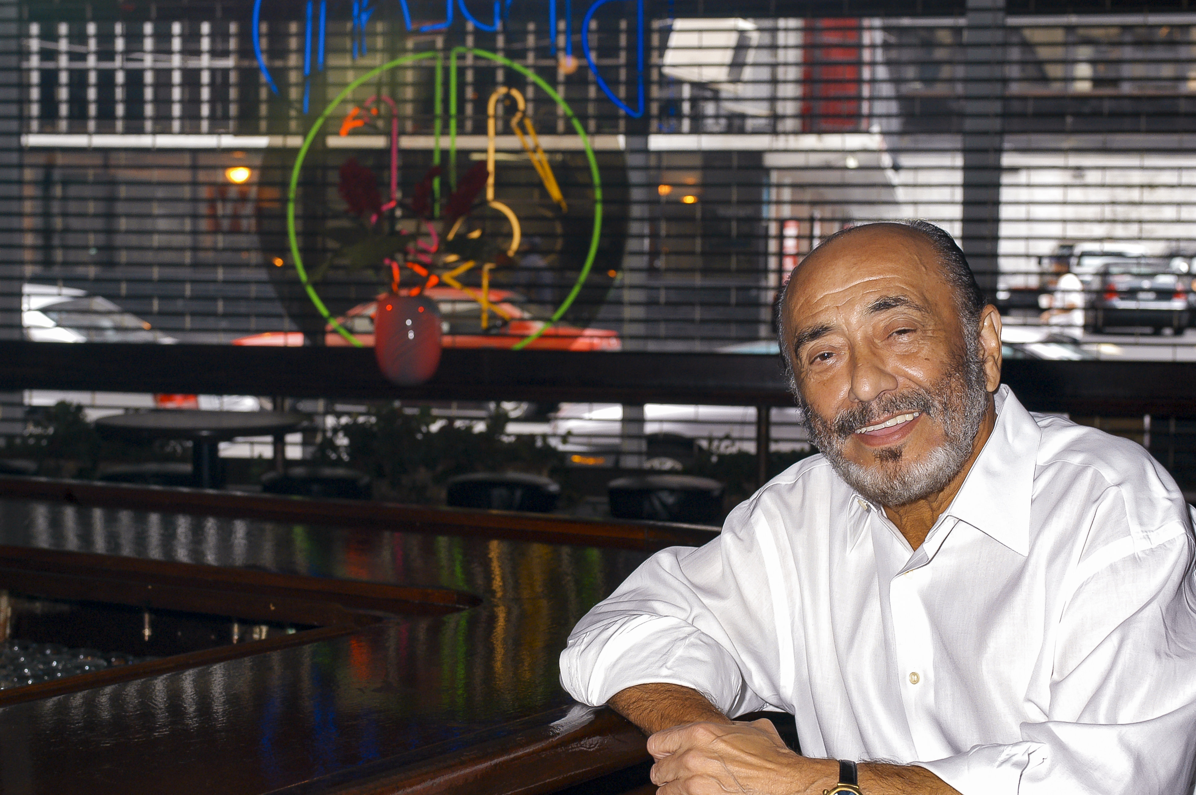 A man in a white shirt leans on a bar counter. A neon sign is visible in the background.