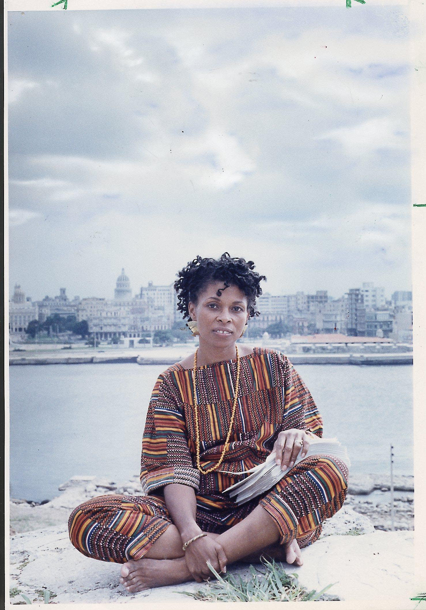 A person sits cross-legged on a rock, wearing a colorful patterned outfit, with a cityscape and water in the background.