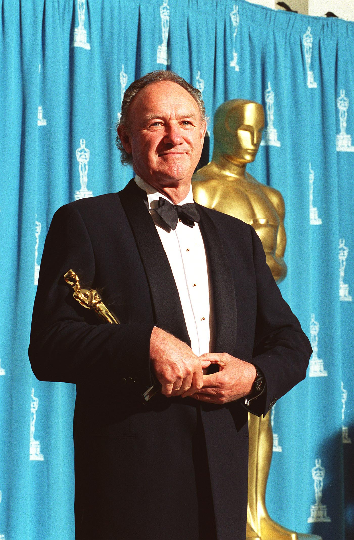 A man in a tuxedo holds an Oscar statue, standing in front of a blue curtain with Oscar logos and a large gold Oscar statue.