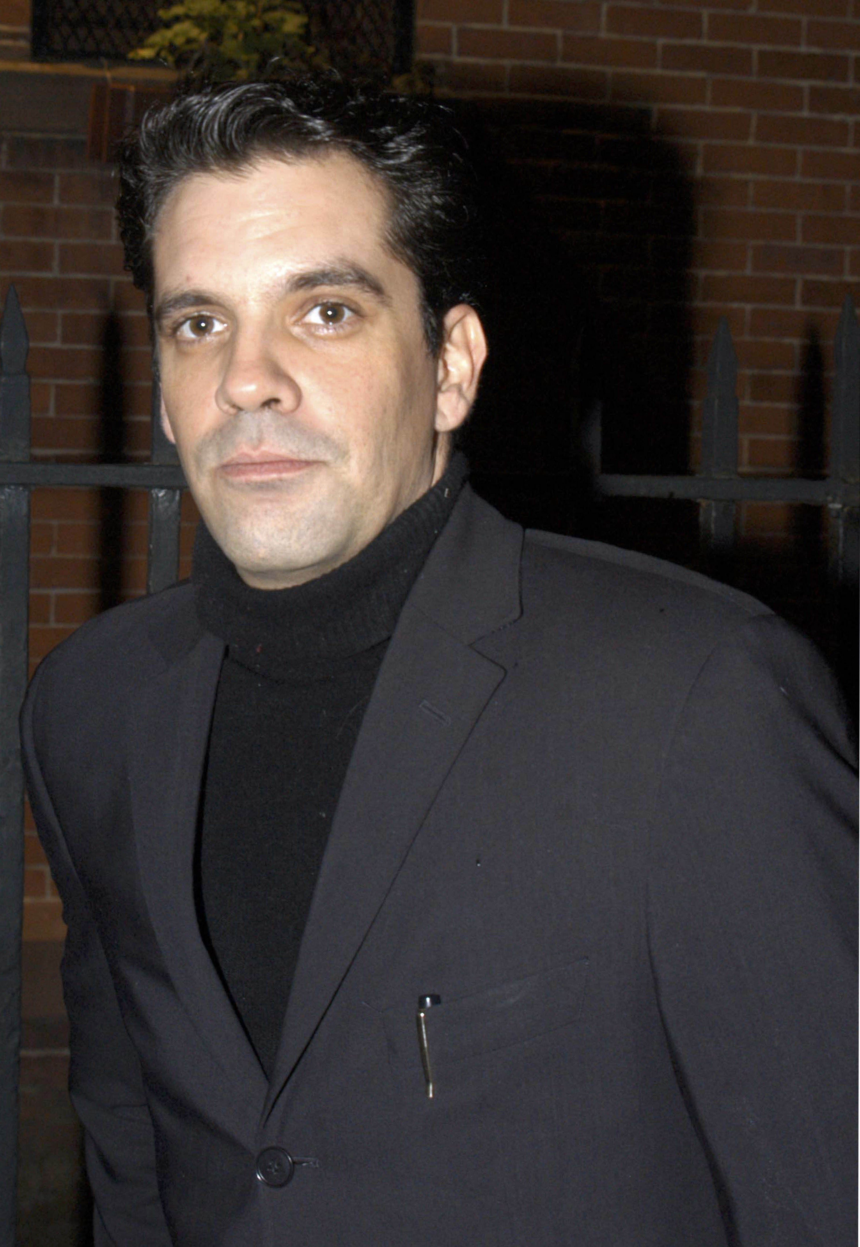 A man in a black turtleneck and blazer stands outdoors at night, with a brick wall and fence in the background.