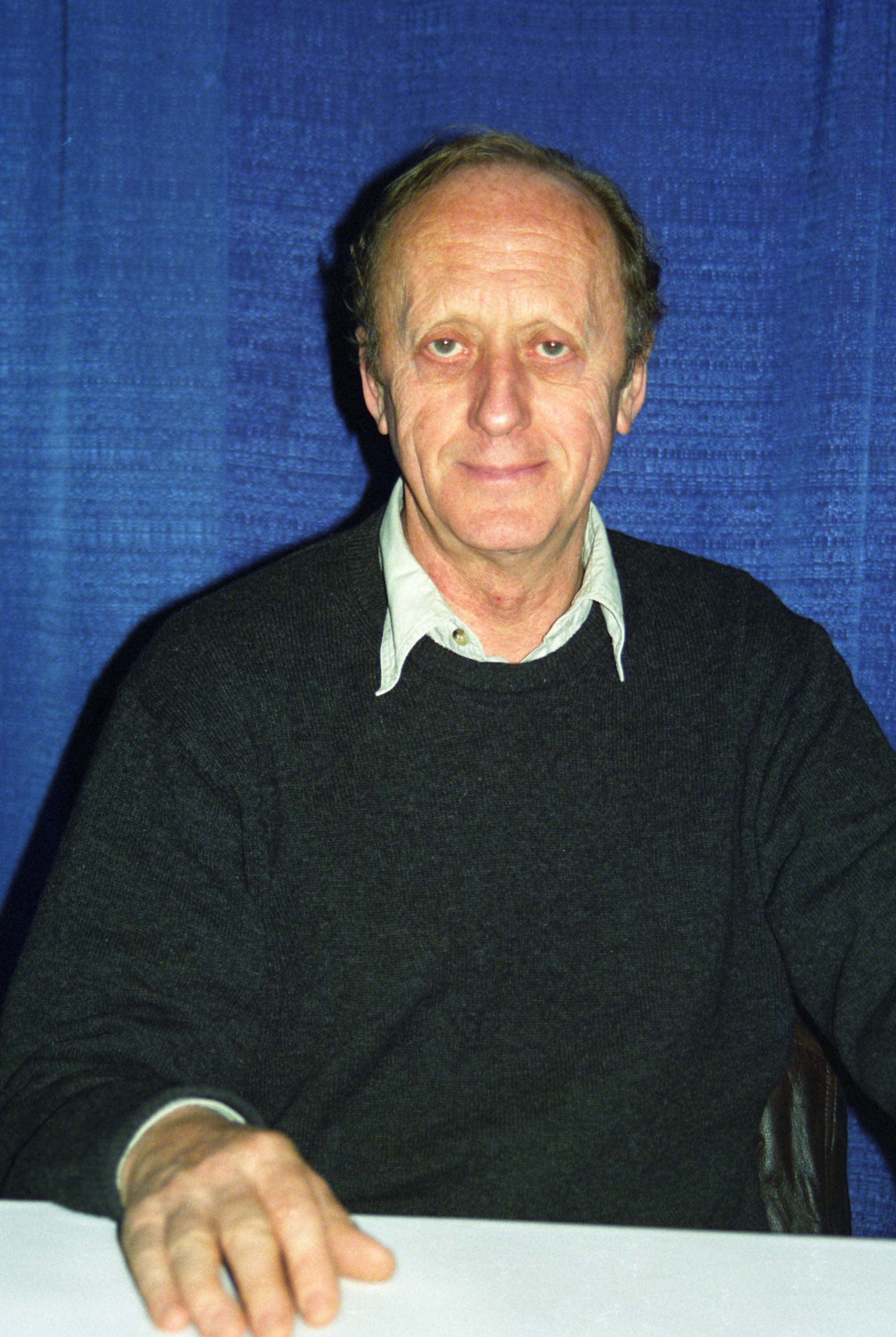 An older man with short hair, wearing a dark sweater over a collared shirt, sitting in front of a blue curtain.
