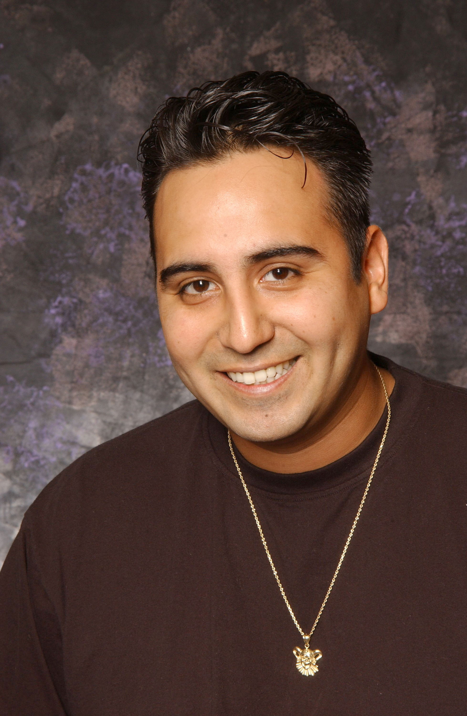 A man with dark hair and a friendly smile, wearing a black shirt and a gold necklace, against a textured dark background.