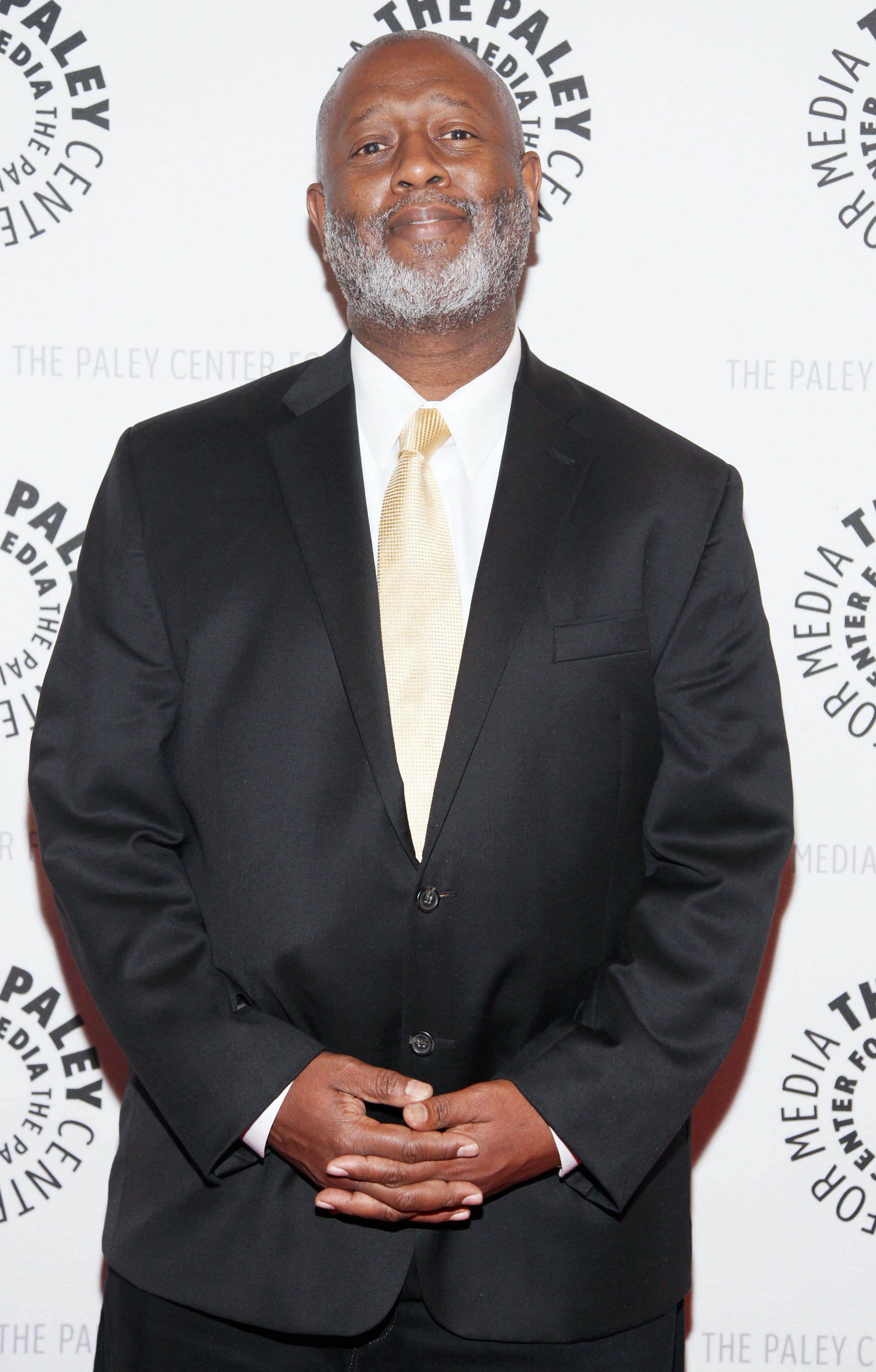 Man in a black suit and yellow tie stands at a Paley Center event, smiling with hands clasped.
