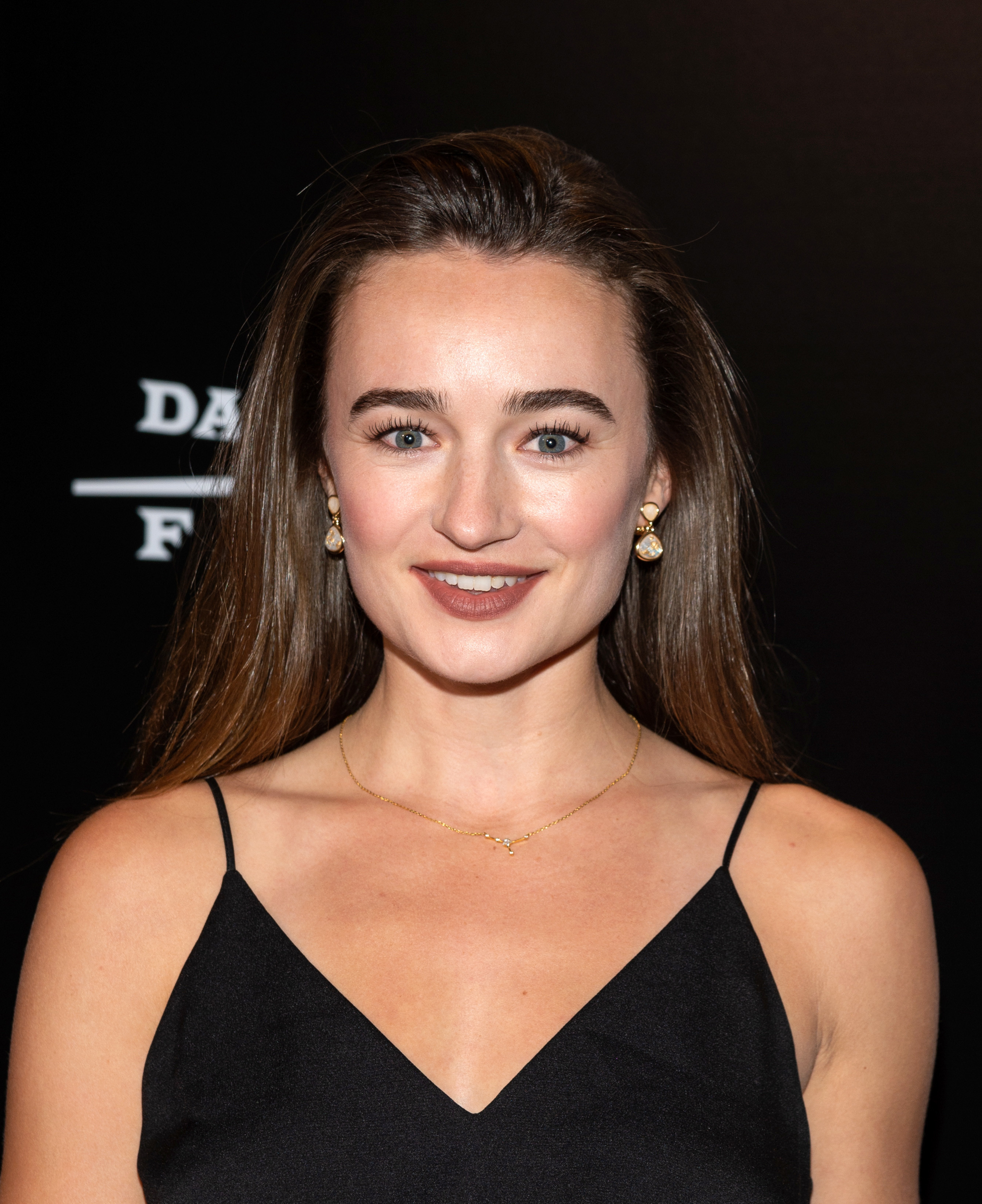 A woman with long brown hair, wearing a black dress and gold earrings, smiles against a dark background.