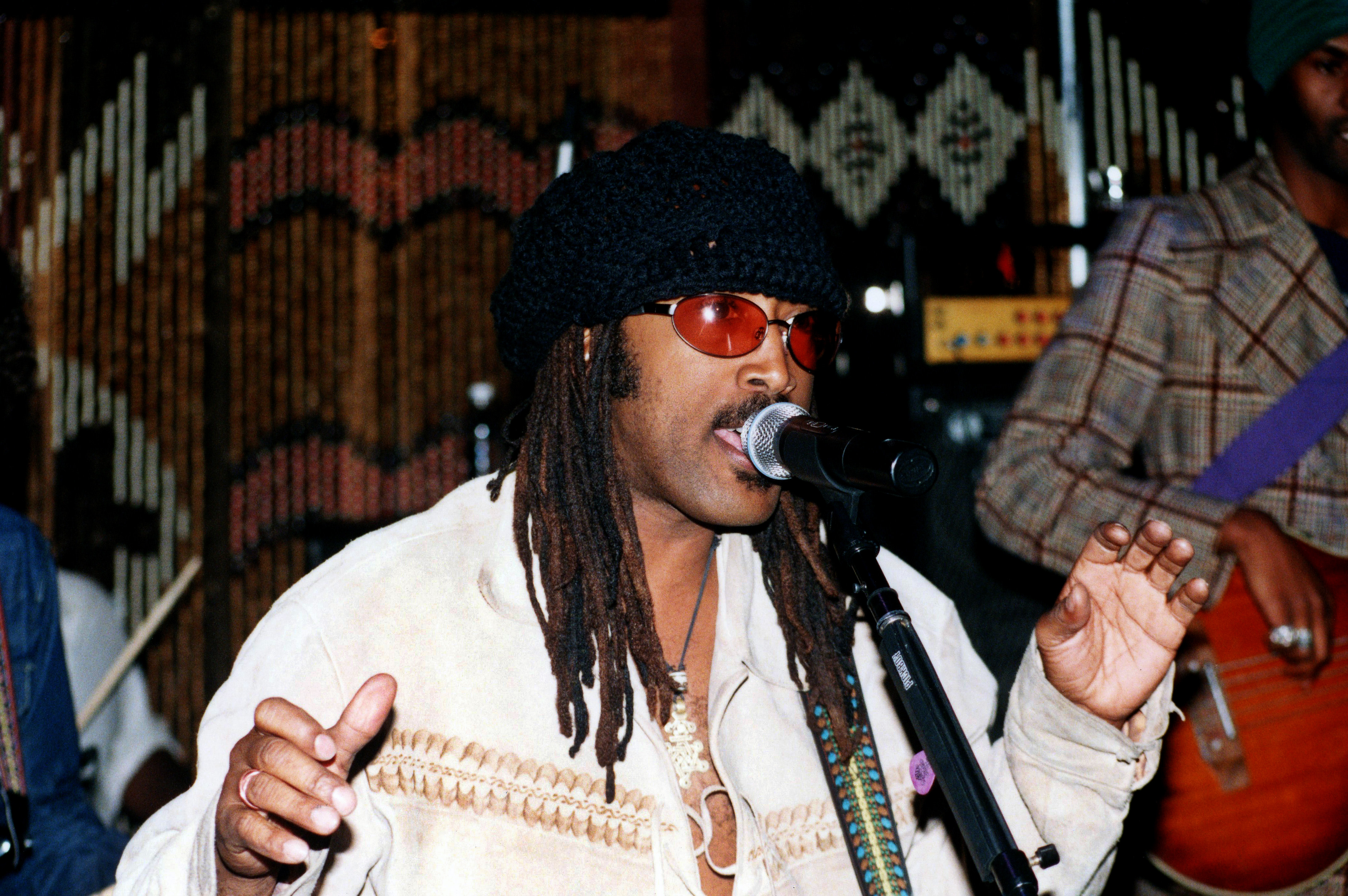 A musician with dreadlocks and sunglasses sings into a microphone, wearing a knitted hat and a light-colored jacket.