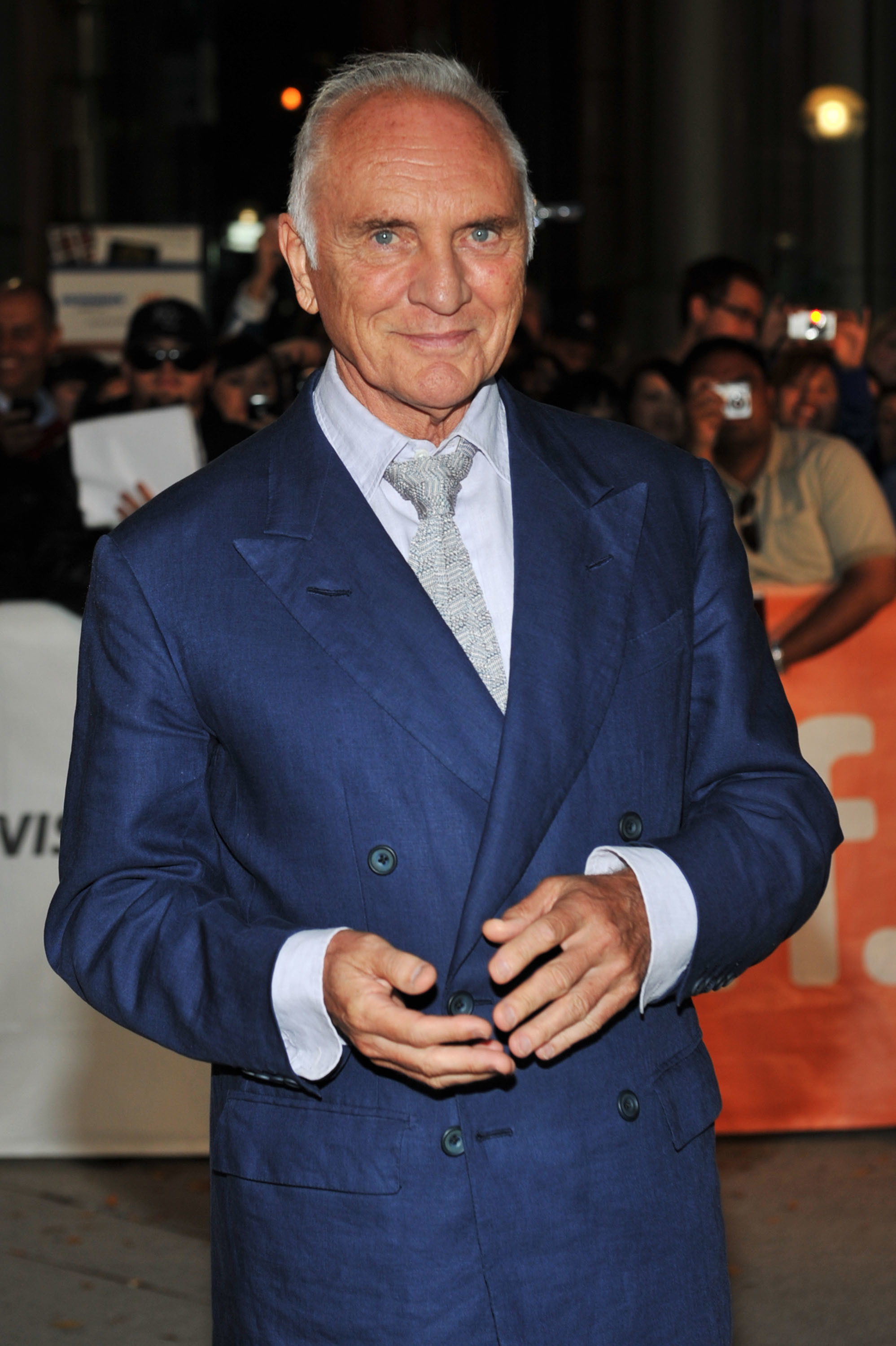 An older man in a blue suit with a light tie, smiling at an event, surrounded by people taking photos.
