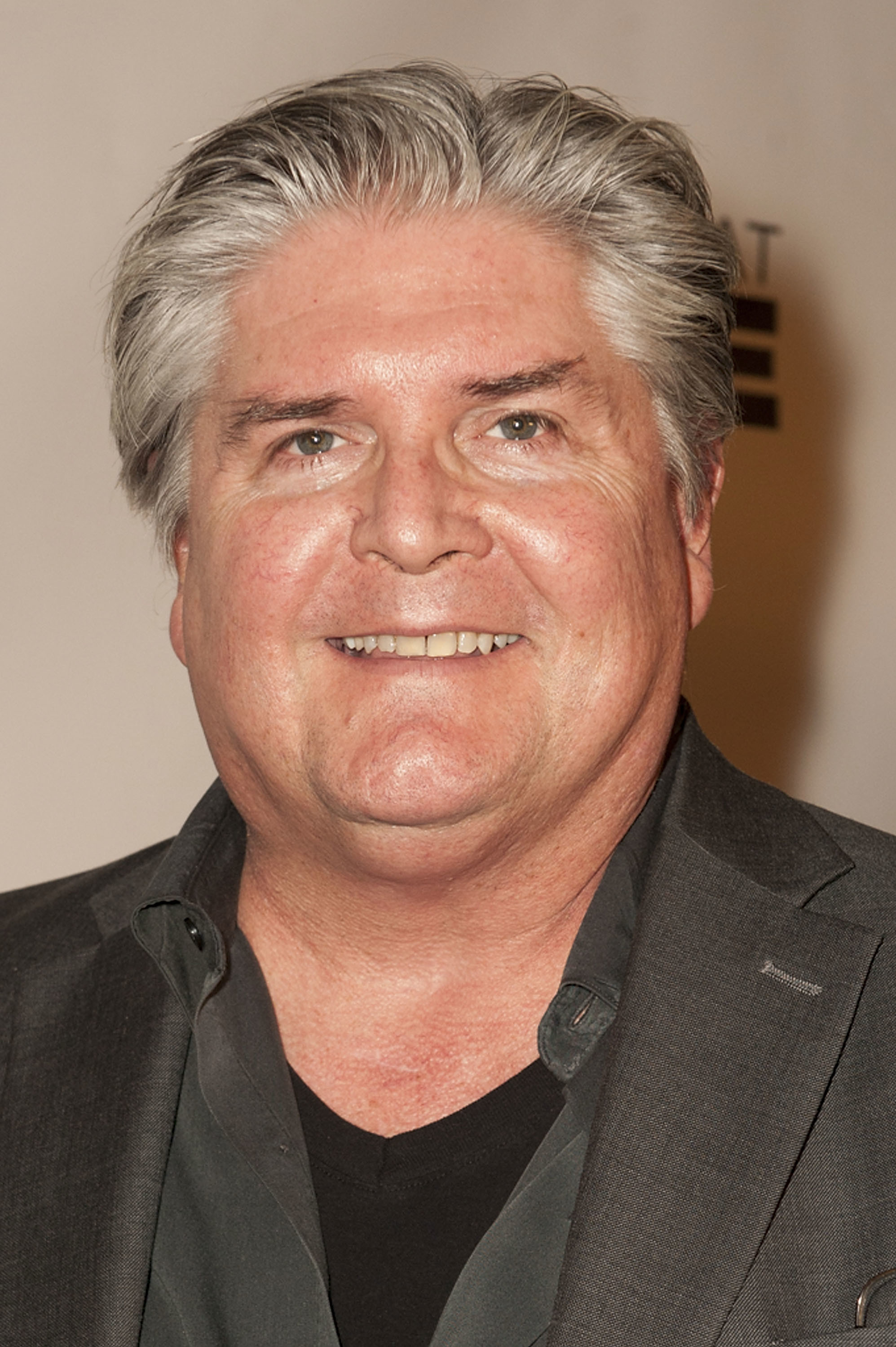A man with gray hair and a black suit smiling at the camera against a neutral background.