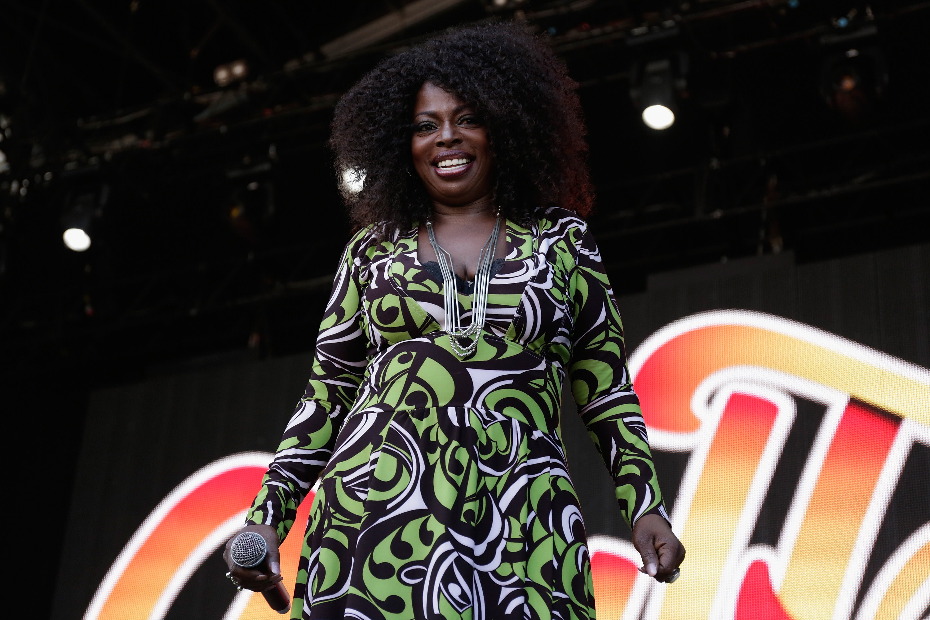 A woman with curly hair performs on stage, wearing a patterned green and black dress and holding a microphone.
