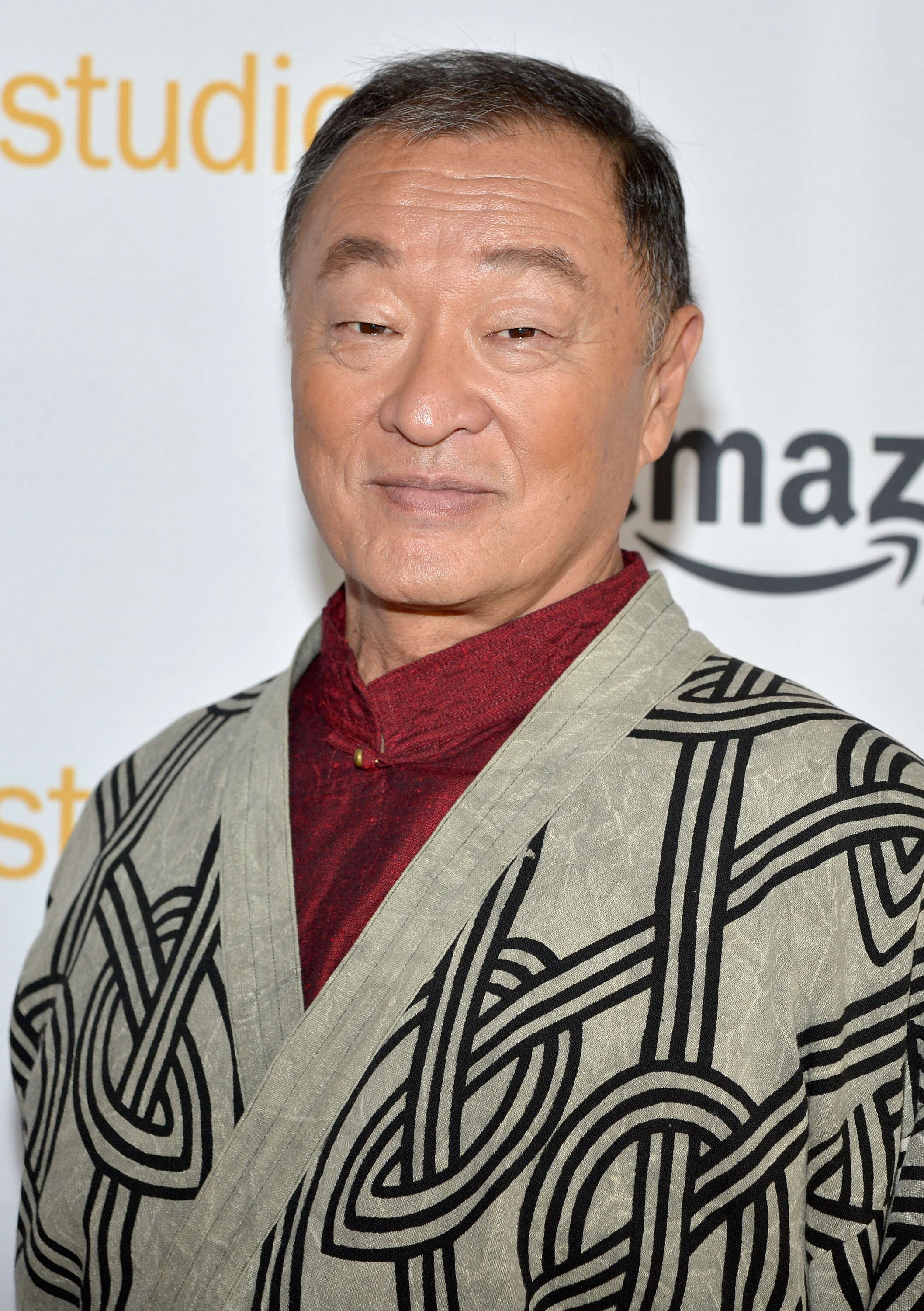 A man in a patterned robe and red shirt at an event with Amazon and Studio logos in the background.