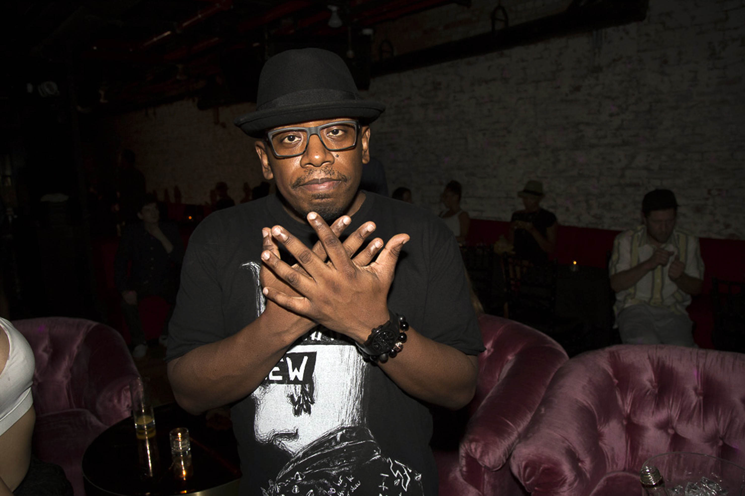 A man wearing a black hat and glasses makes a hand gesture, standing in a dimly lit room with pink chairs and a brick wall.