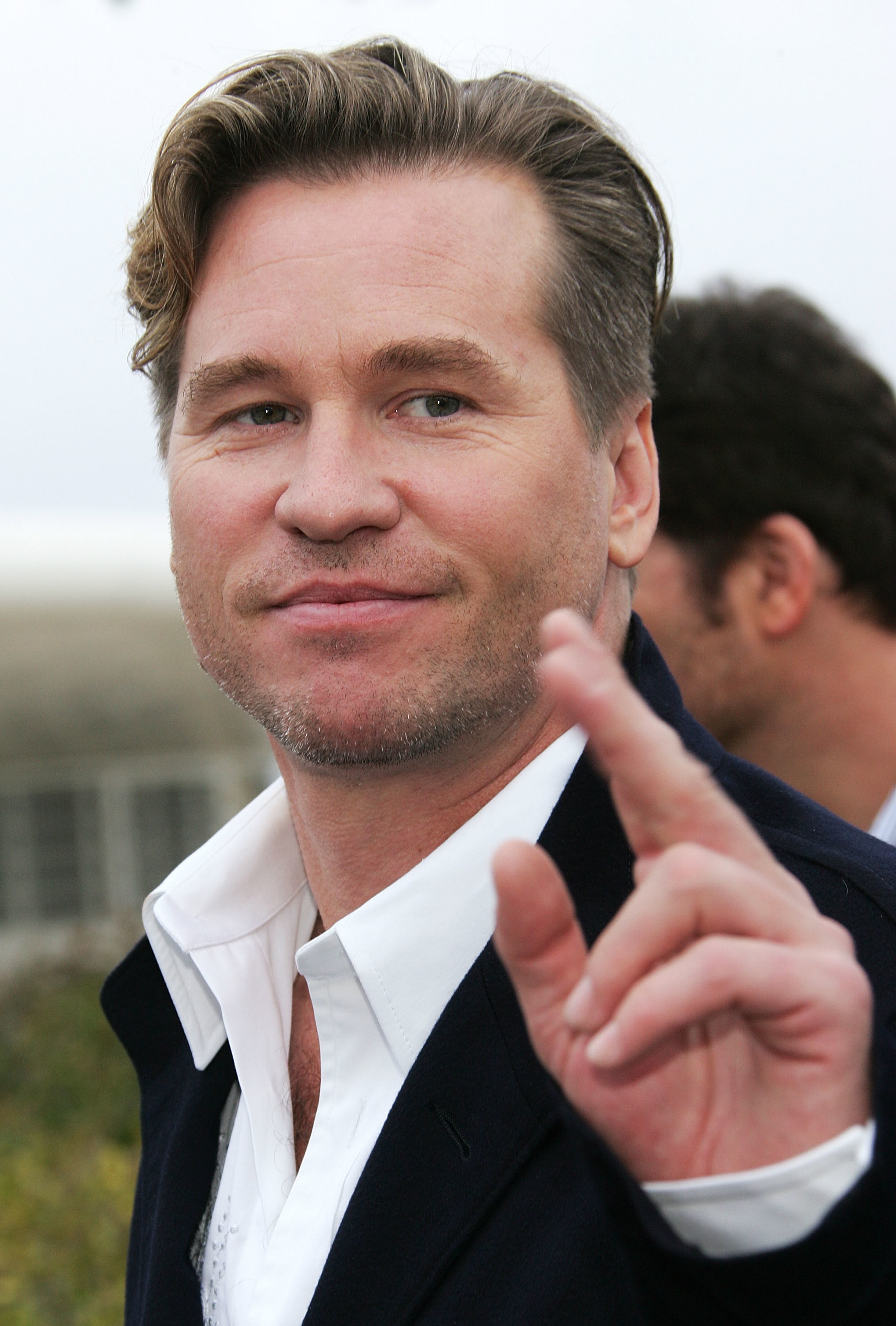A man with light brown hair and a slight smile gestures towards the camera, wearing a white shirt and dark jacket.
