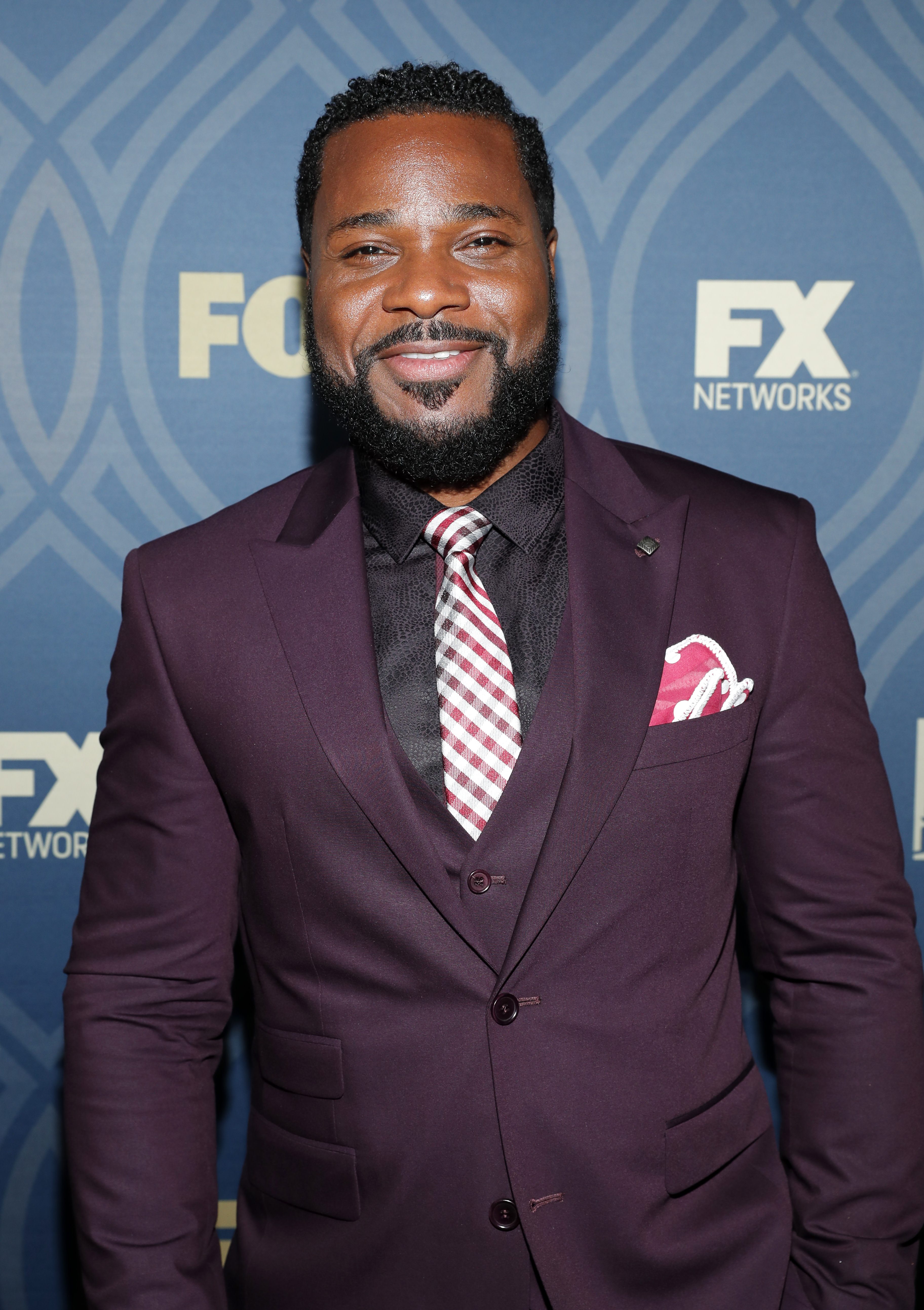 Man in a maroon suit with a checkered tie, standing against a backdrop with FX Networks logos.