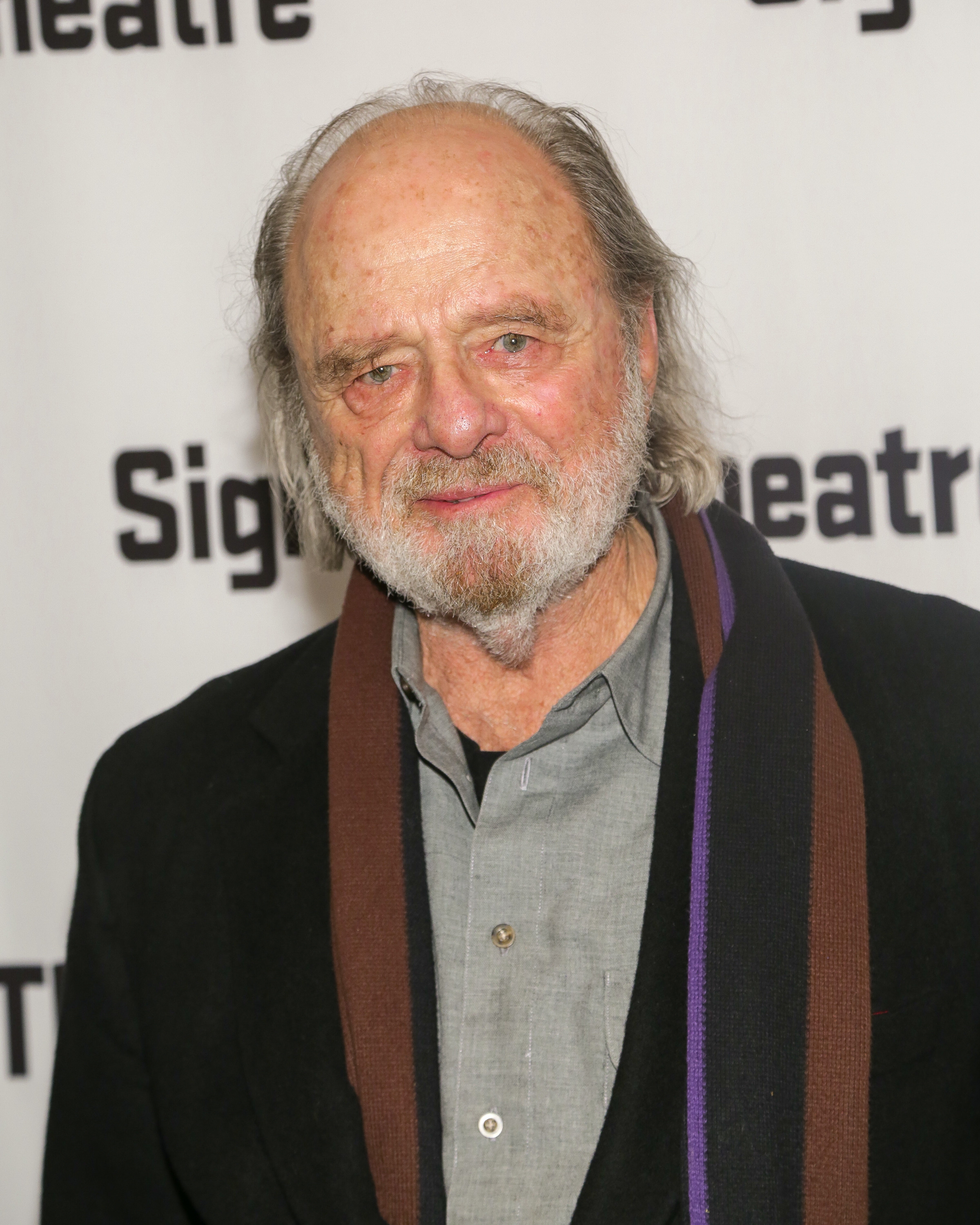 An older man with a beard and long hair, wearing a gray shirt, black jacket, and scarf, stands in front of a