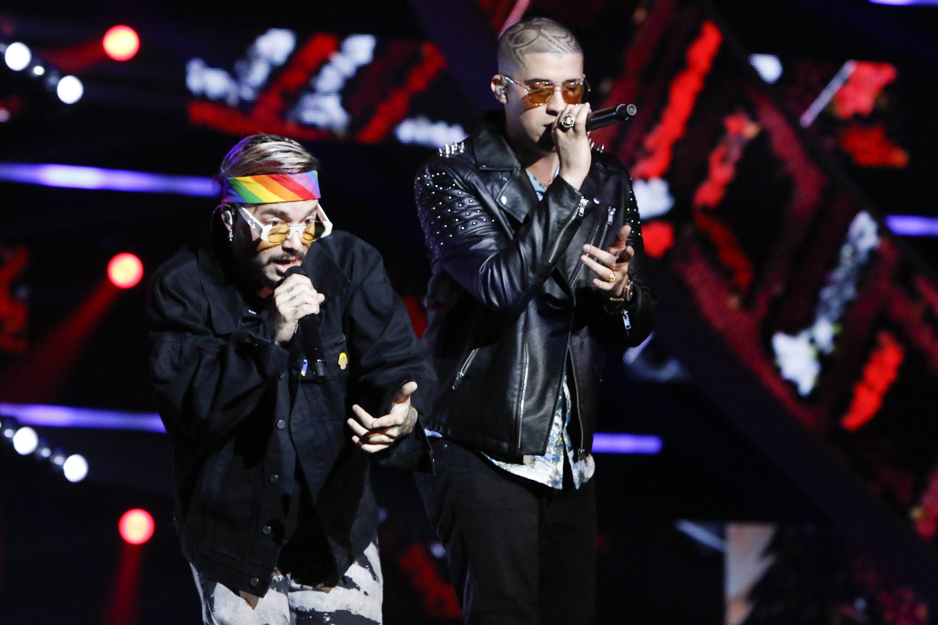 Two performers on stage, one with a rainbow headband and yellow glasses, and the other in a studded leather jacket, both singing.