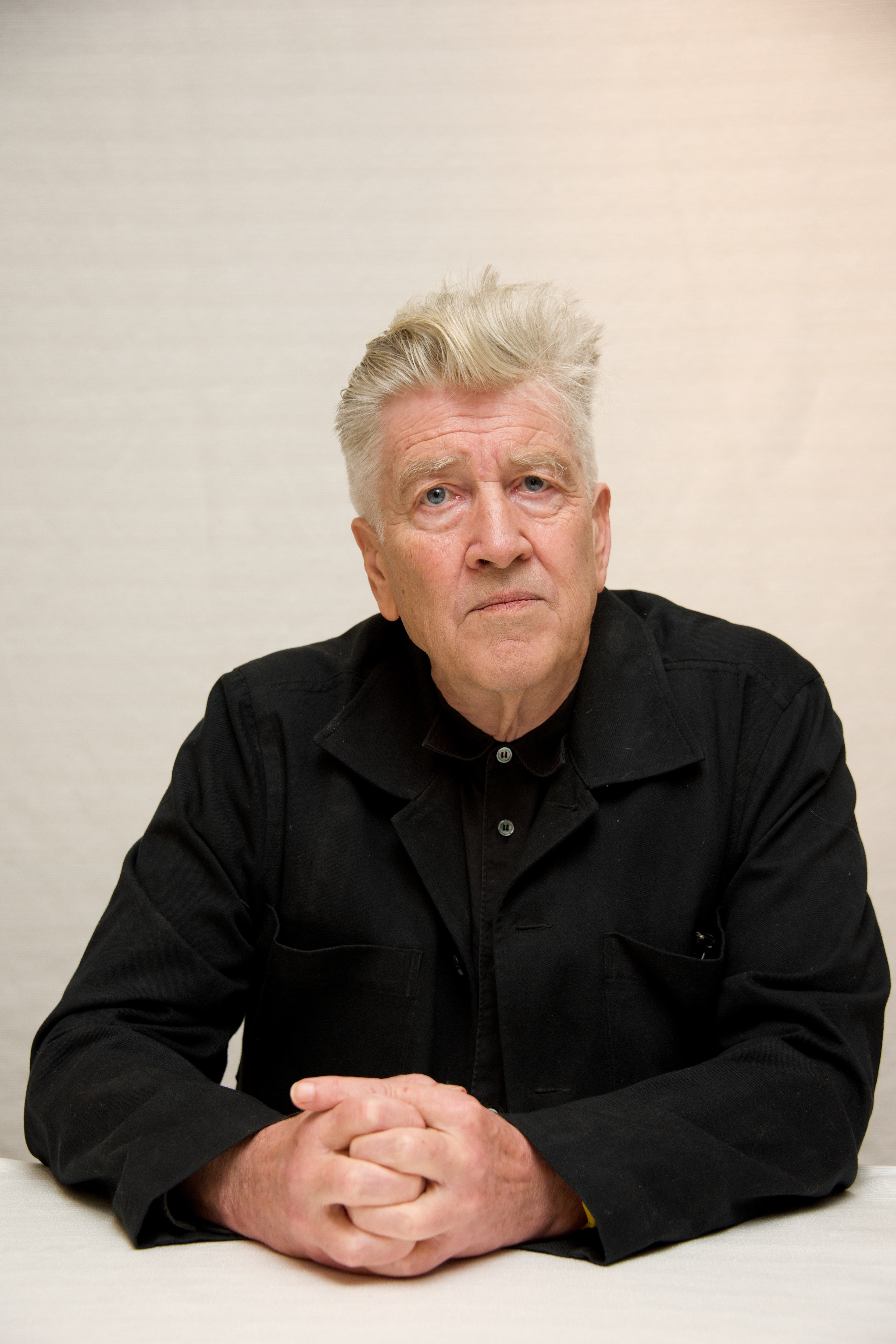 A man with white hair and a serious expression, wearing a black jacket, sits with hands clasped on a table.