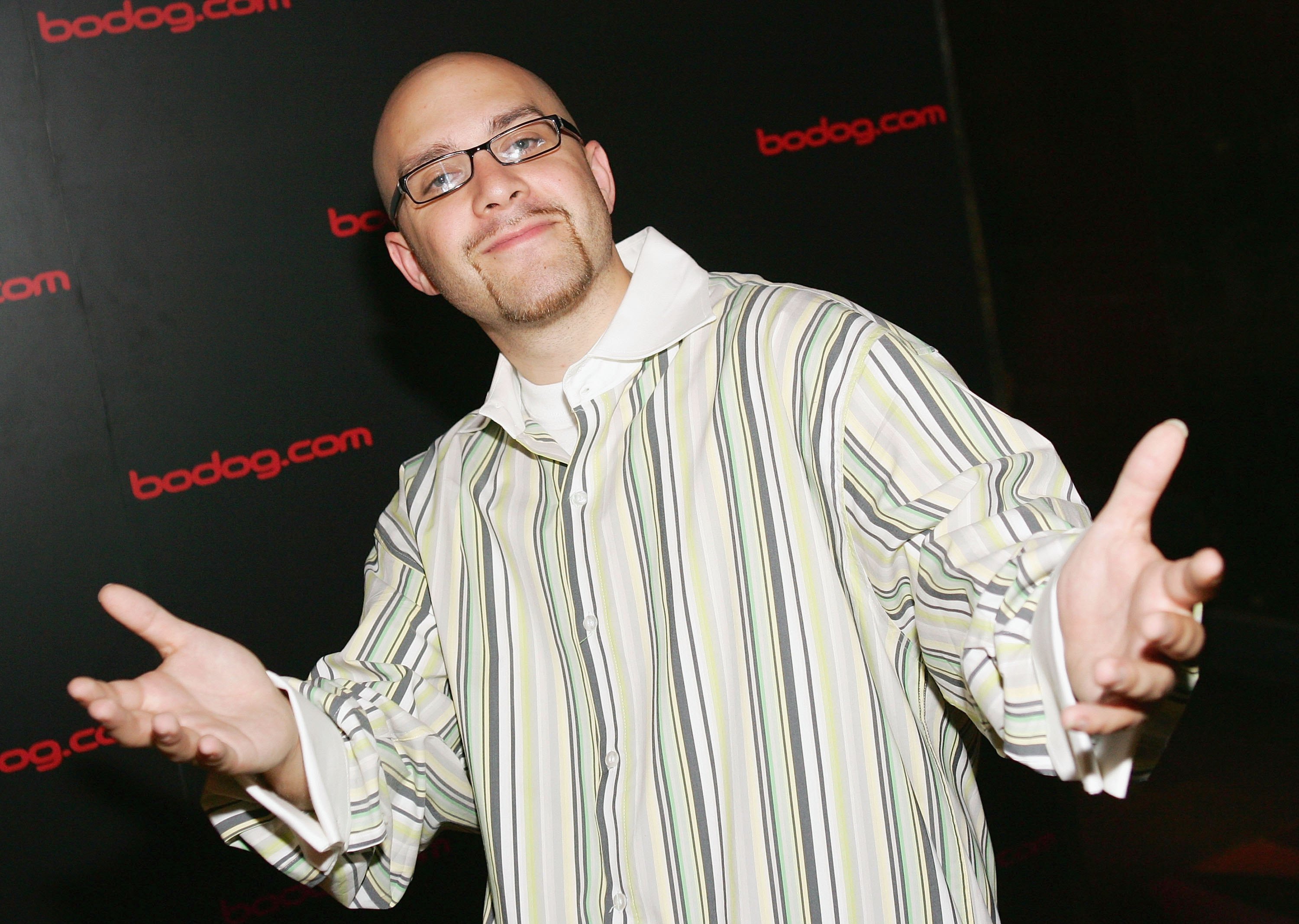 A man with glasses and a goatee poses with open arms in front of a backdrop with 