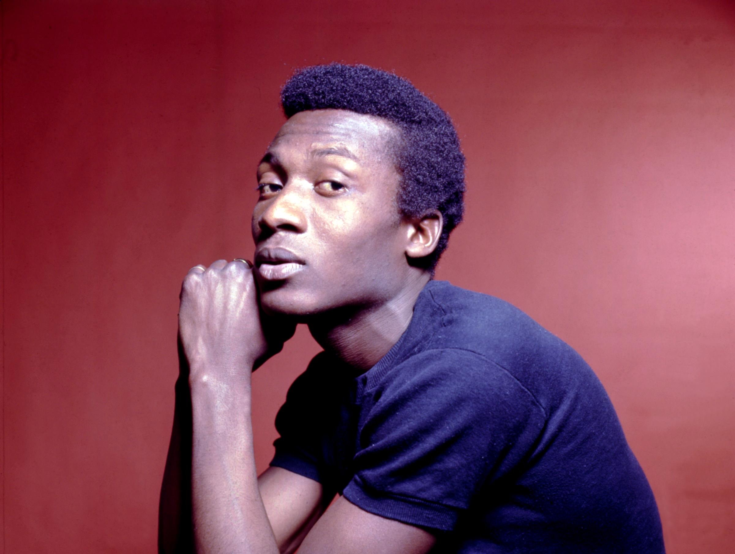 A person in a contemplative pose against a red background, wearing a dark shirt, with short hair.