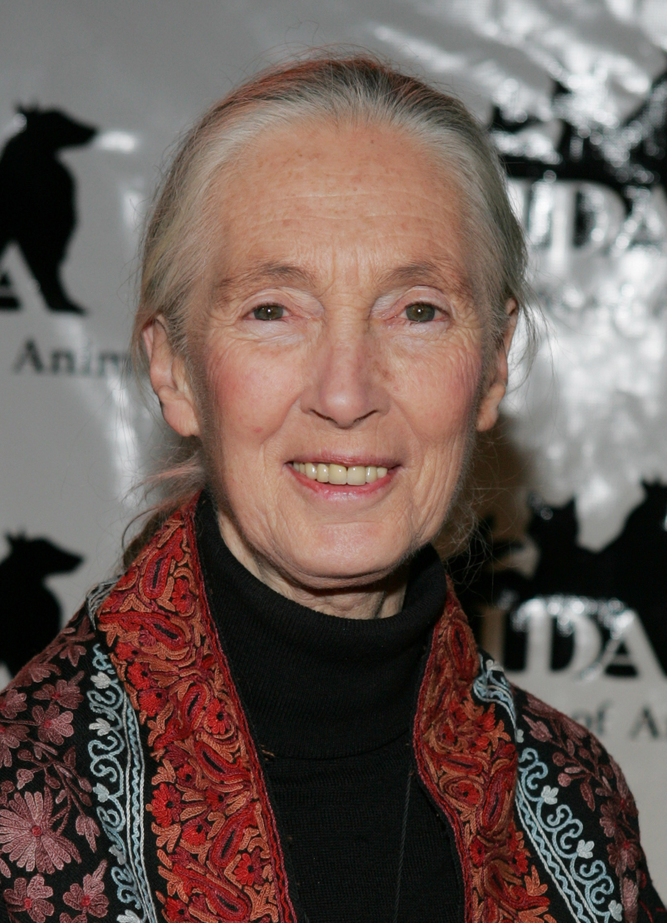 An elderly woman with long gray hair, wearing a black top and a red patterned shawl, smiles at the camera.
