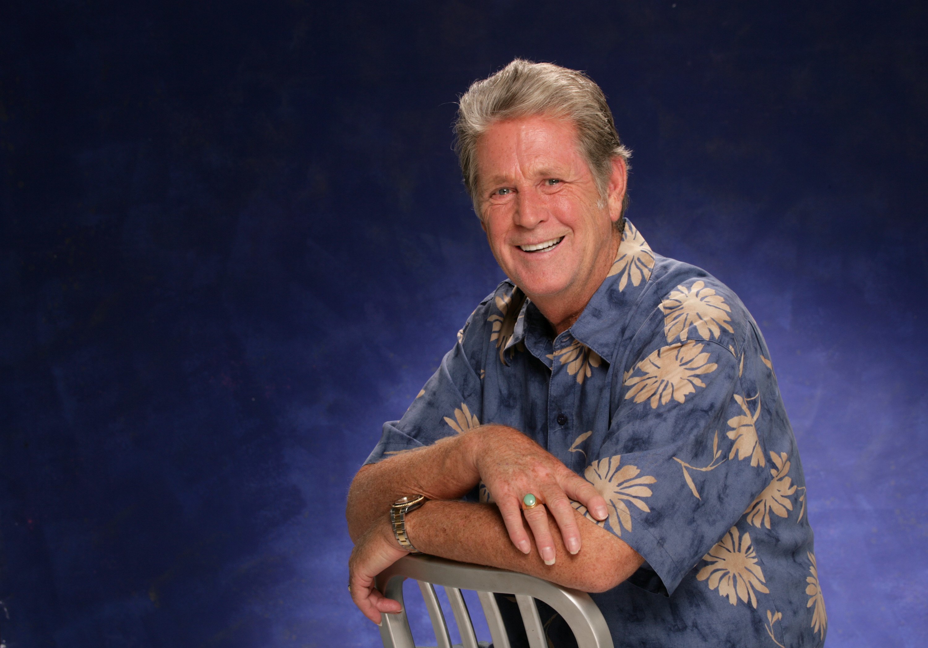 A smiling man in a floral shirt leans on a chair against a blue background.