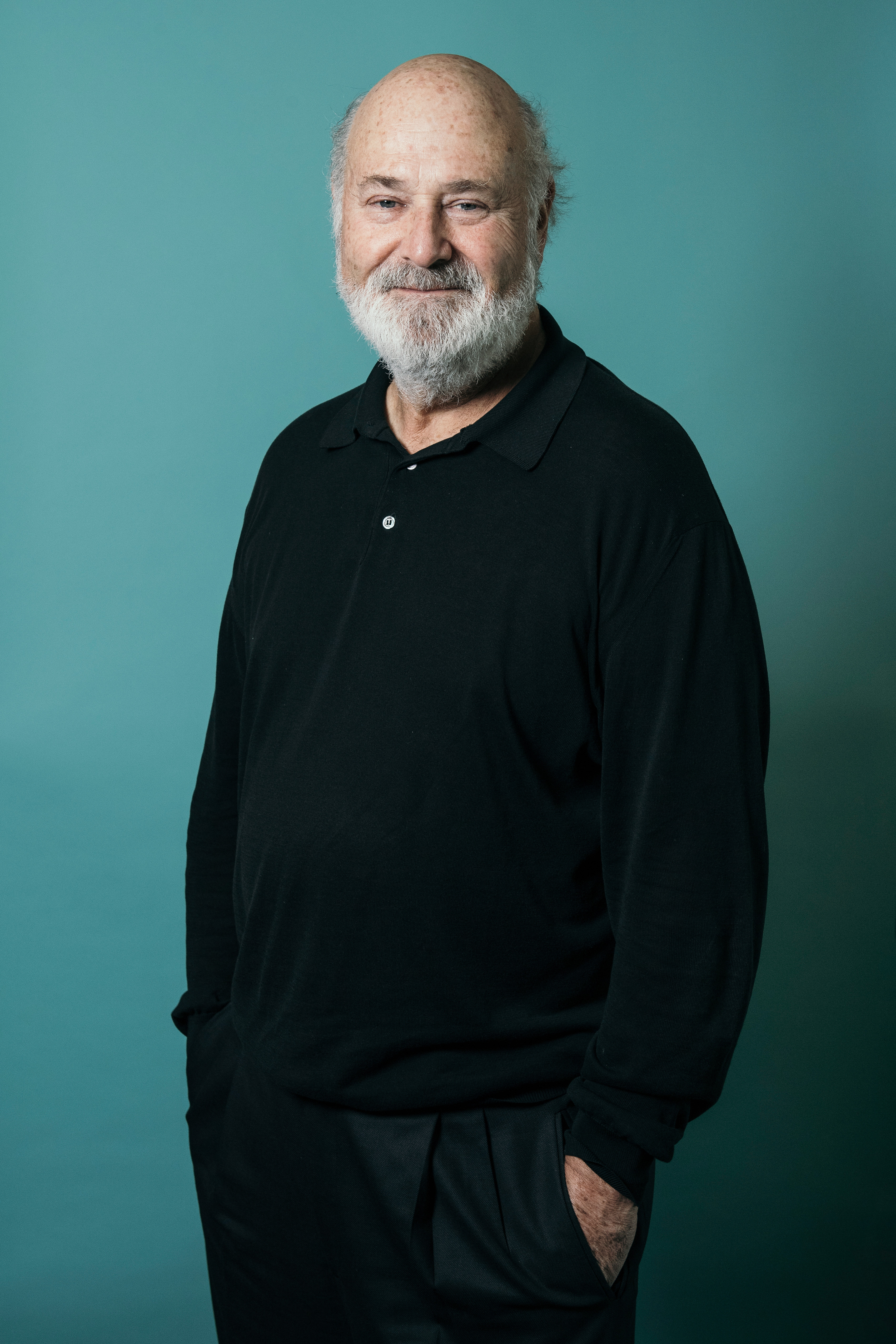 An older man with a white beard, wearing a black shirt, stands against a teal background.