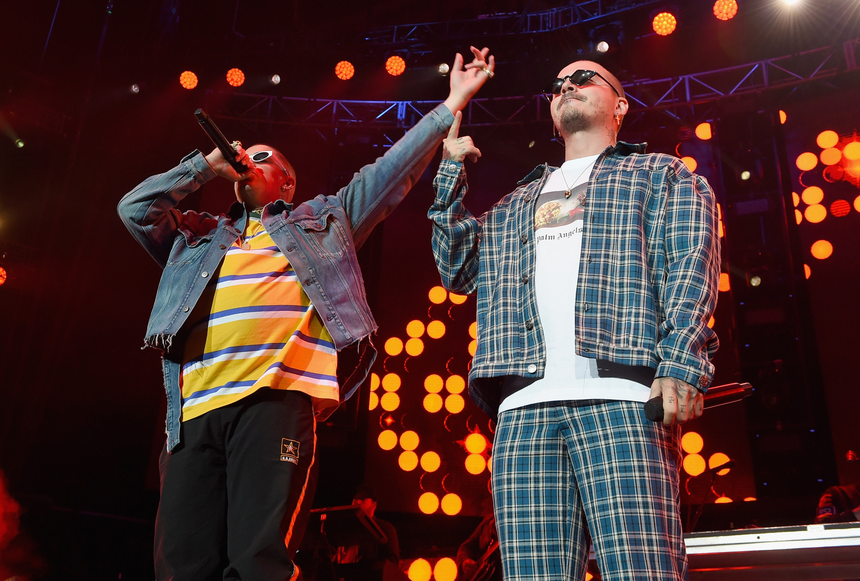 Two performers on stage, one in a striped shirt and denim jacket, the other in plaid, both wearing sunglasses, with vibrant lights behind.