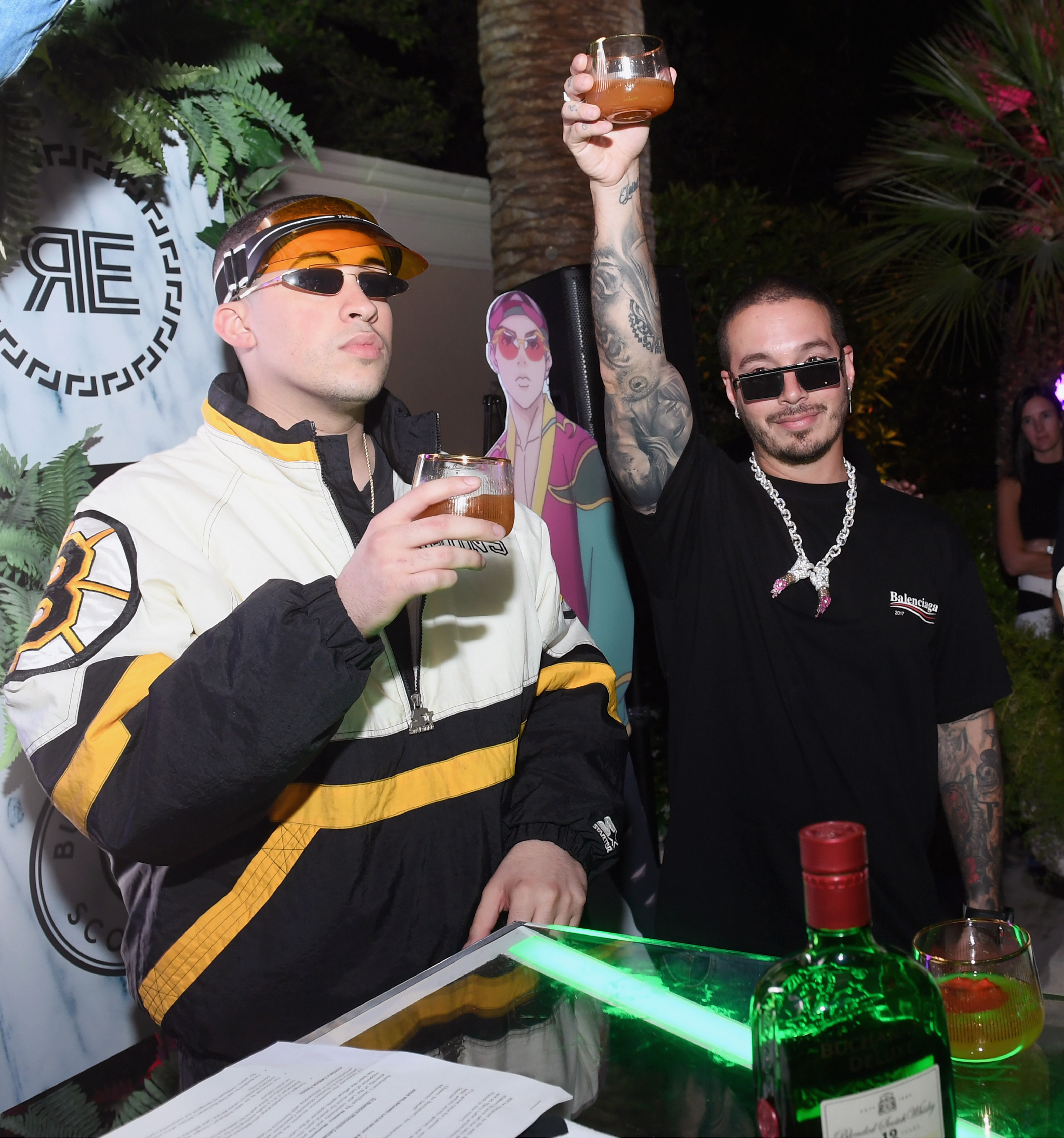 Two men wearing sunglasses raise drinks at an outdoor event, surrounded by greenery and a decorative backdrop.
