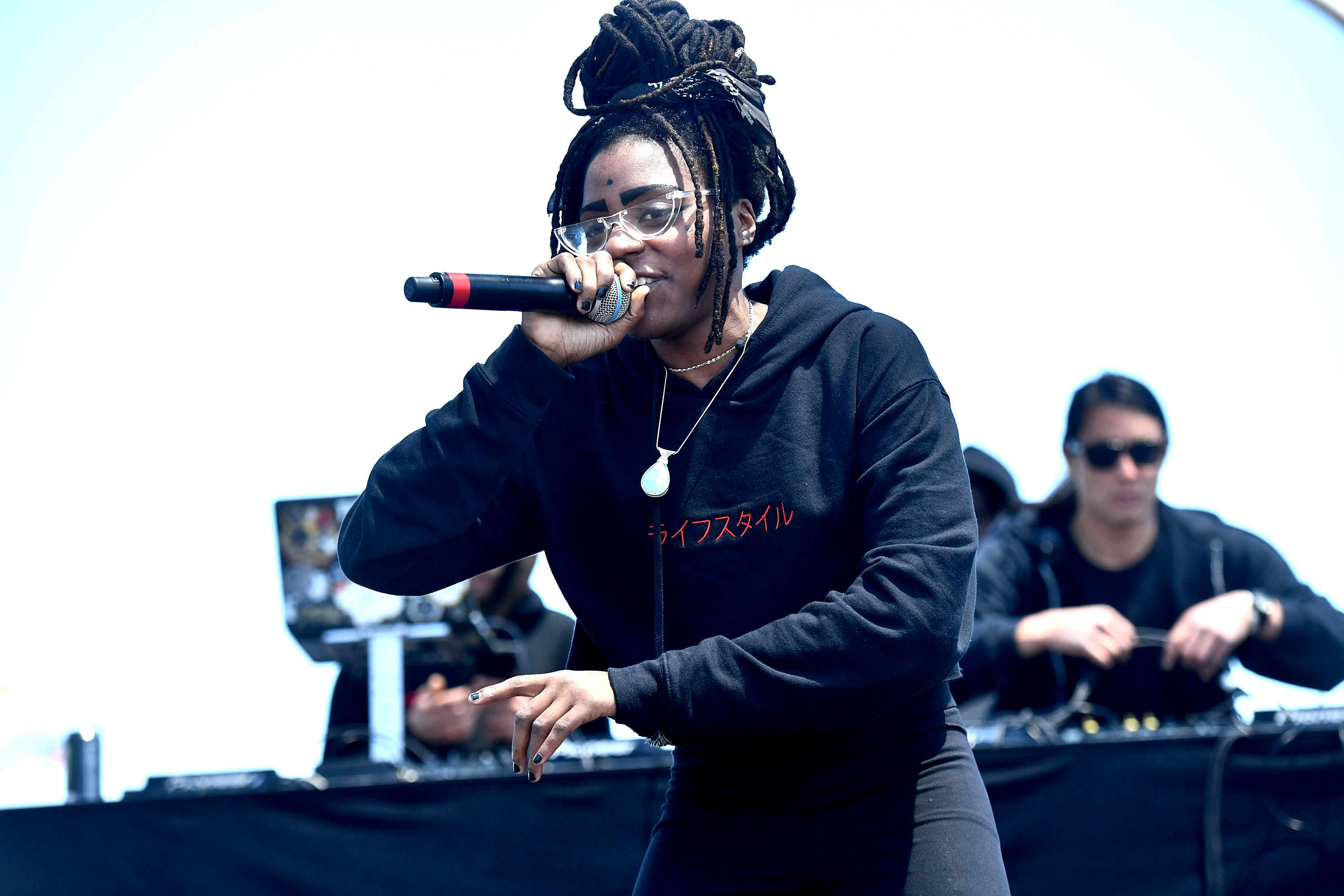 Chelsea Reject with long braids and glasses raps into a microphone on stage, wearing a black hoodie. DJ in the background.