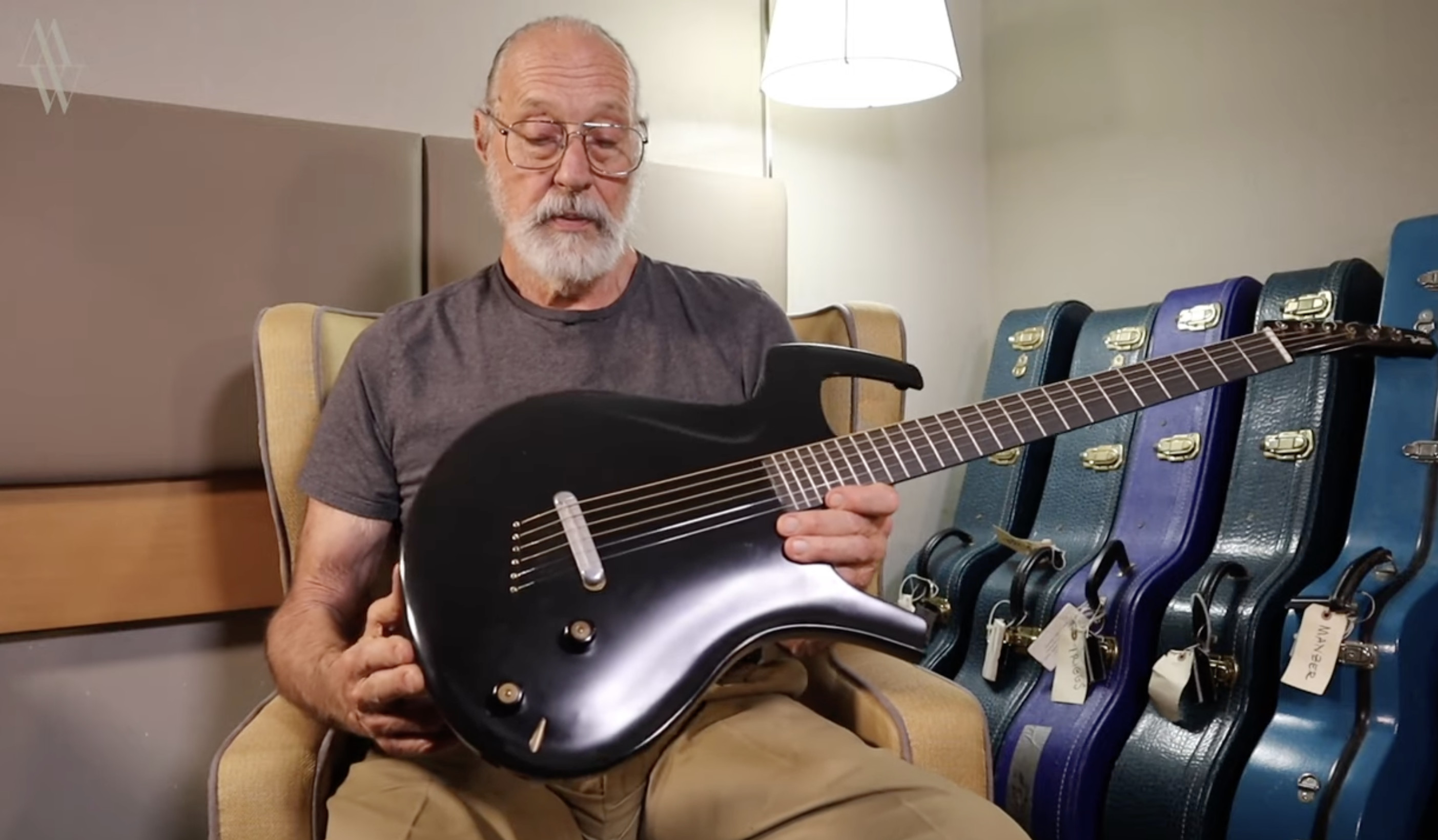 An older man with glasses and a beard sits in a chair holding a sleek, black guitar. Several guitar cases are visible in the background.