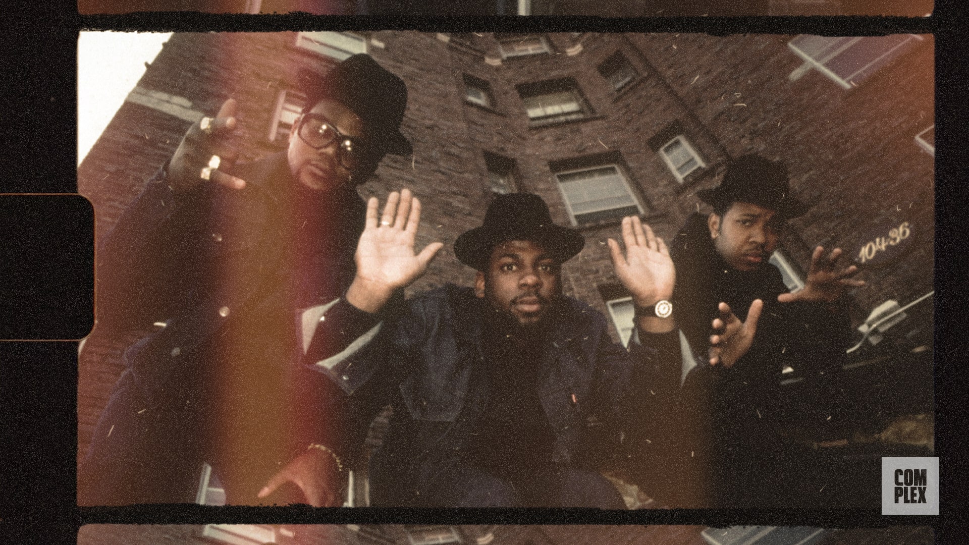 Three men in black hats and jackets pose with hands raised in front of a brick building, exuding an urban, vintage style.