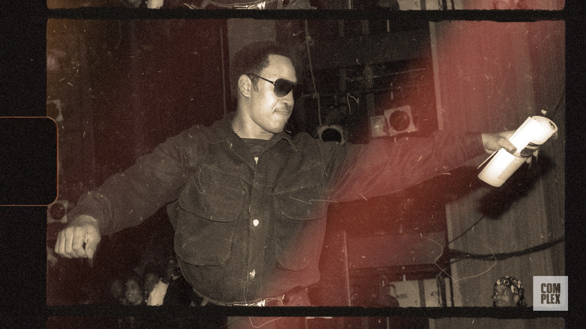 A person in sunglasses and a dark jacket energetically holds a microphone on stage, with a vintage filter applied.