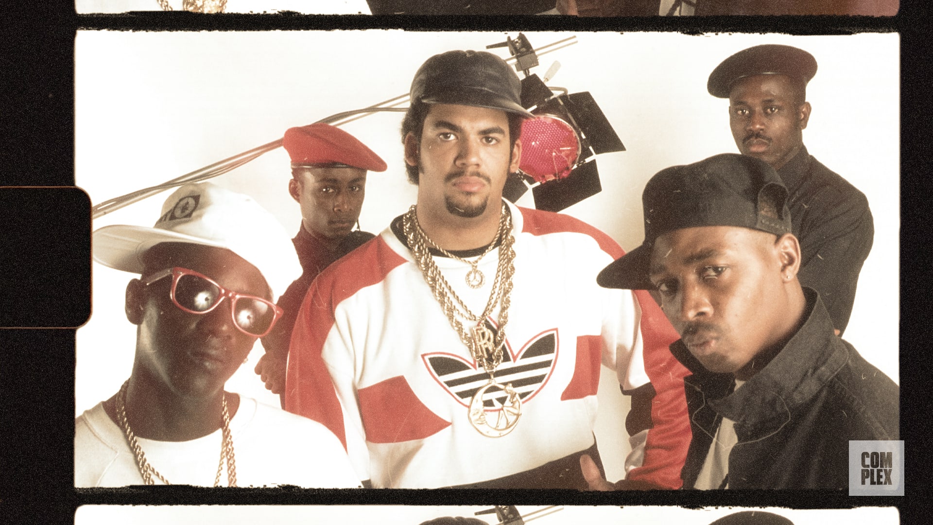 A group of five men, including Flavor Flav and Chuck D from Public Enemy, posing in hip-hop attire with hats and chains.