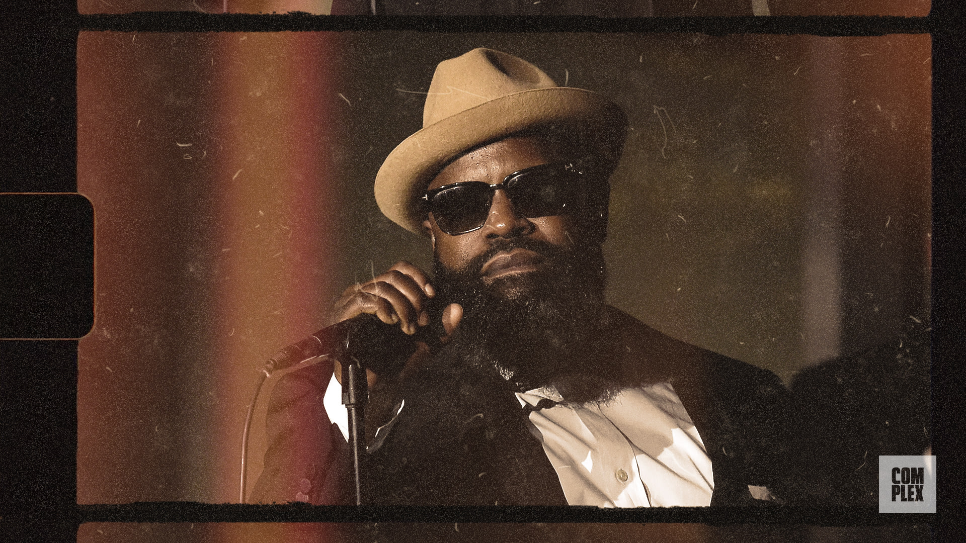 A man wearing sunglasses and a hat holds a microphone, dressed in a suit. The image has a vintage film effect with a 