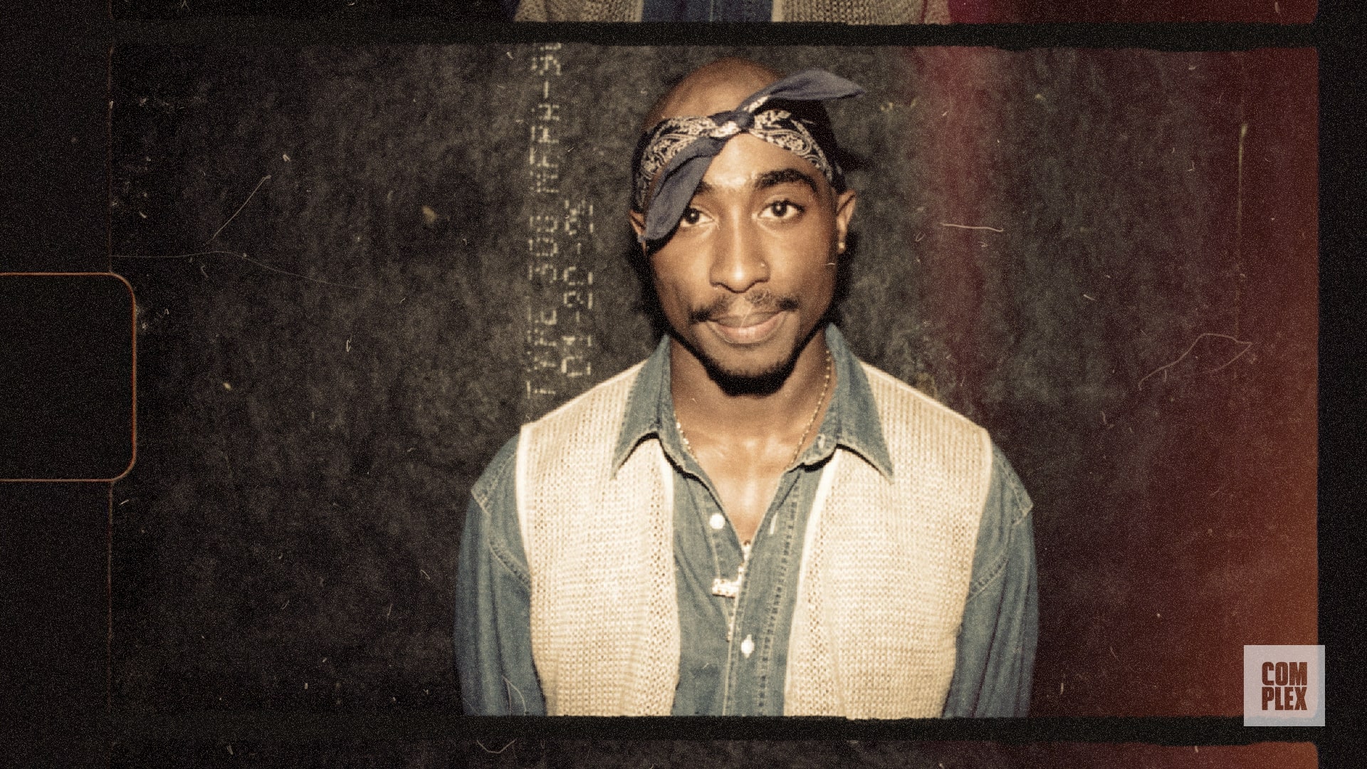 Tupac Shakur wearing a denim shirt and beige vest, with a blue bandana tied around his head, standing against a textured background.