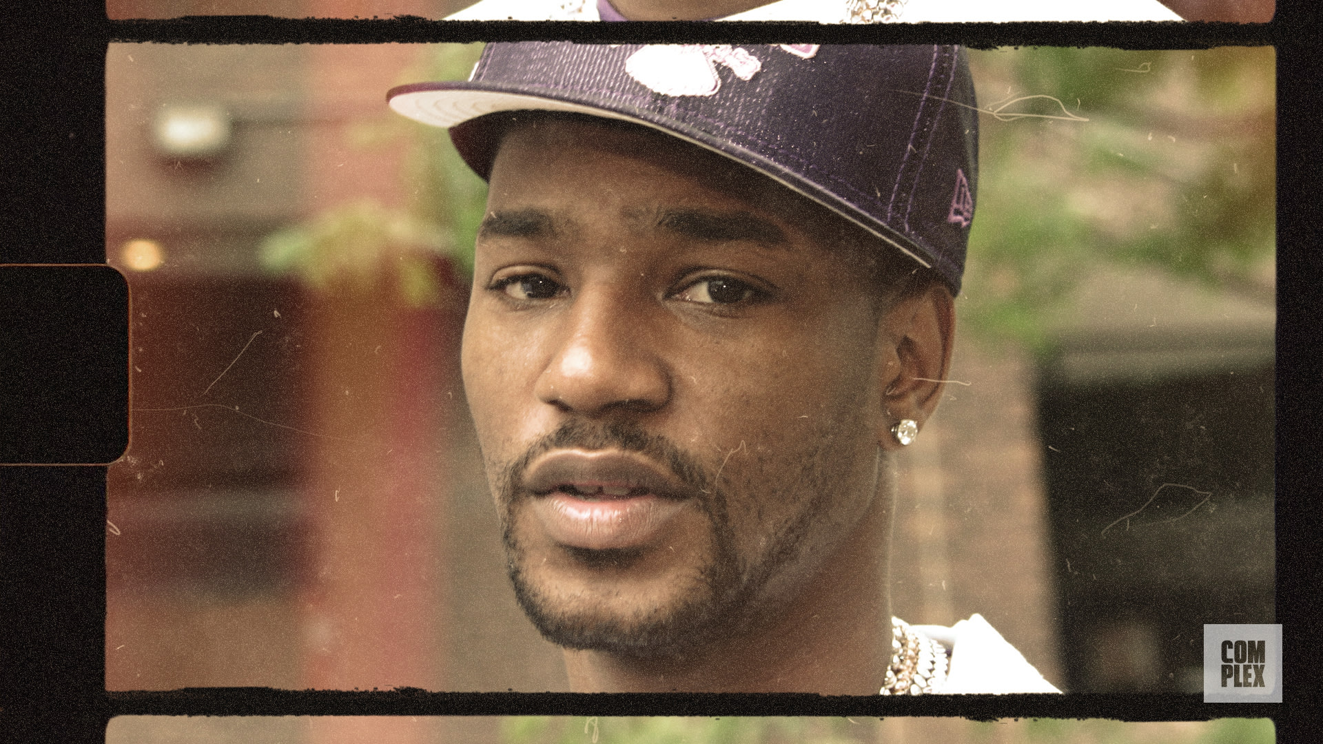 A man wearing a purple cap and earrings, with a thoughtful expression, in an outdoor setting.