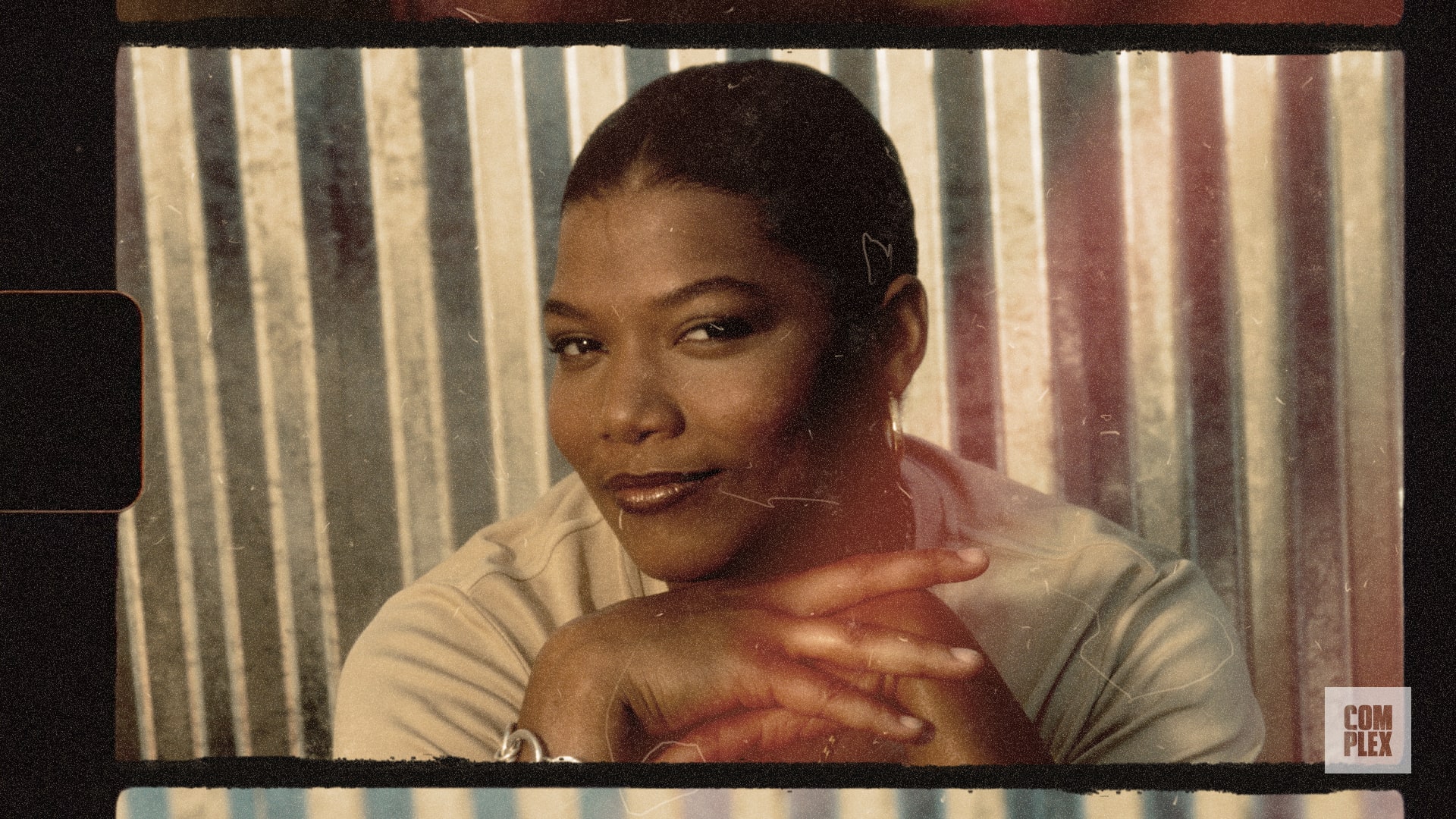 A person with short hair, smiling, rests their chin on their hands against a corrugated metal background.
