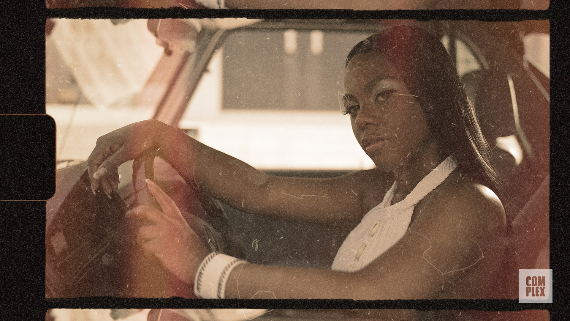 A woman with long hair sits confidently in a vintage car, wearing a white top. The image has a retro, sepia-toned effect.