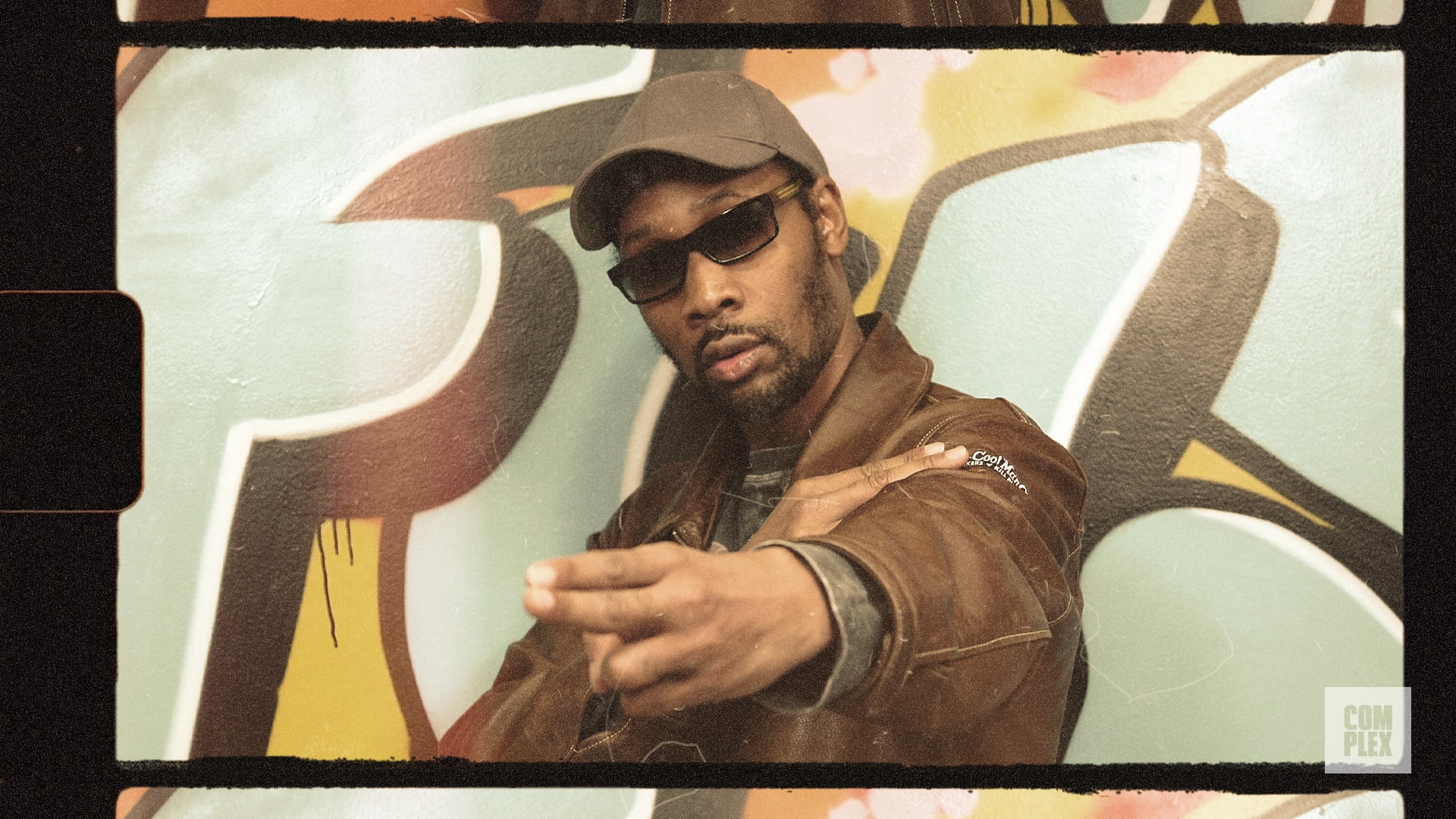 A man wearing sunglasses and a cap poses in front of graffiti, pointing with his hand. The image has a vintage film-like border.