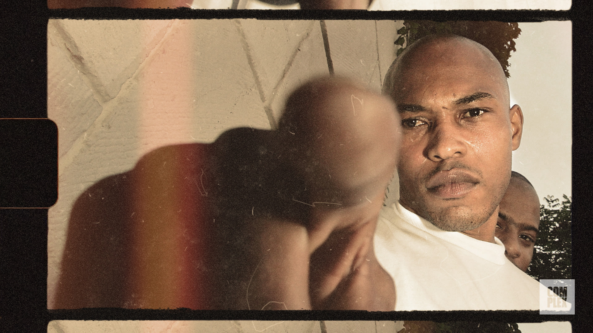 A man in a white shirt points towards the camera with a serious expression. Another person is partially visible behind him.