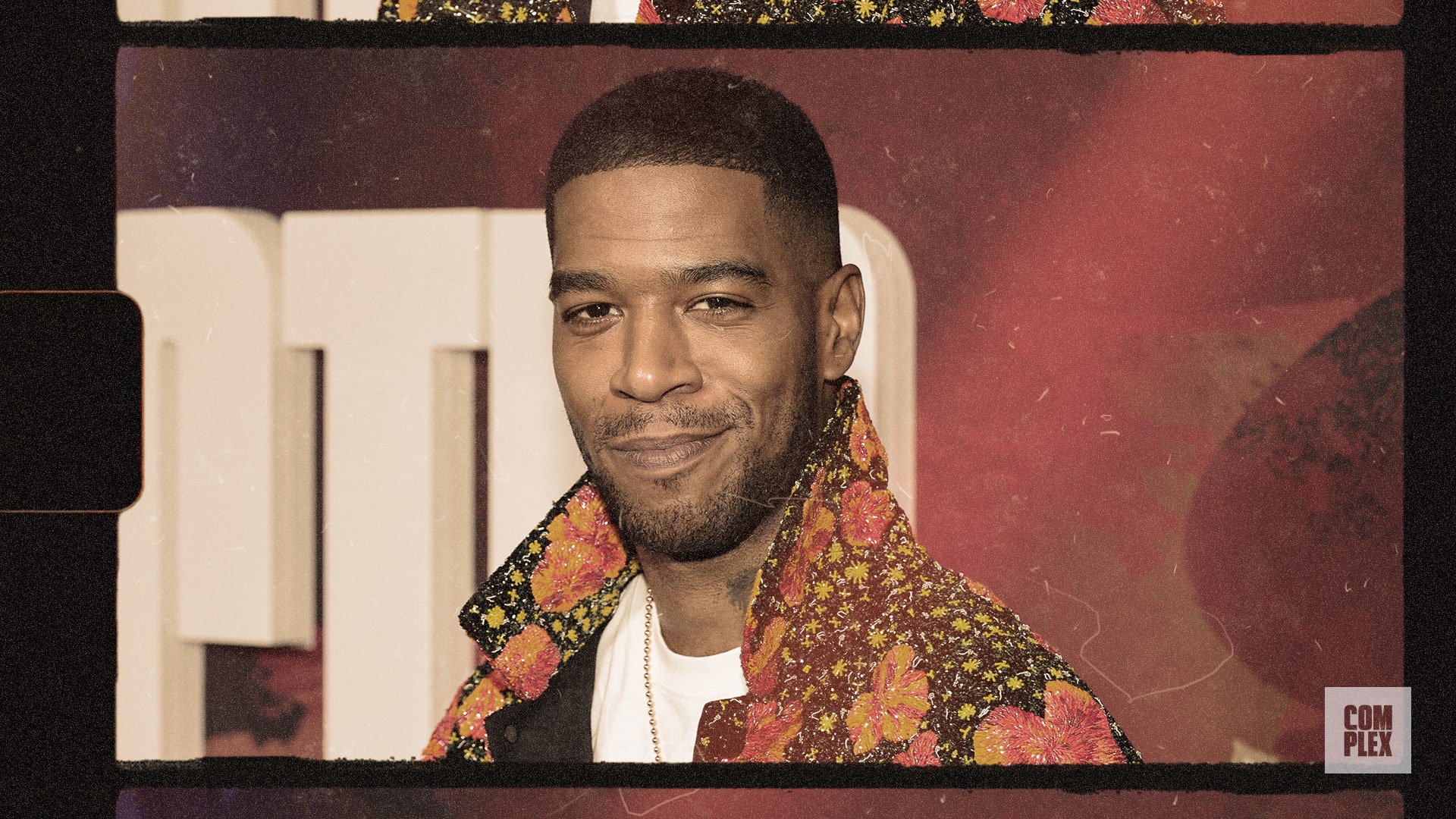 Kid Cudi, wearing a floral jacket, smiles at an event with a red and white background.