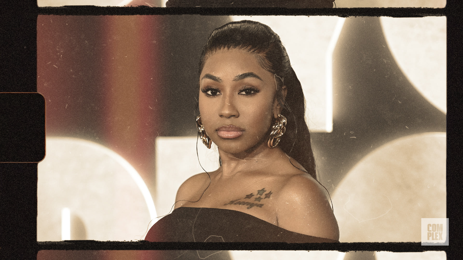 A woman with long hair and hoop earrings looks at the camera. She wears a black outfit and has a tattoo on her shoulder.