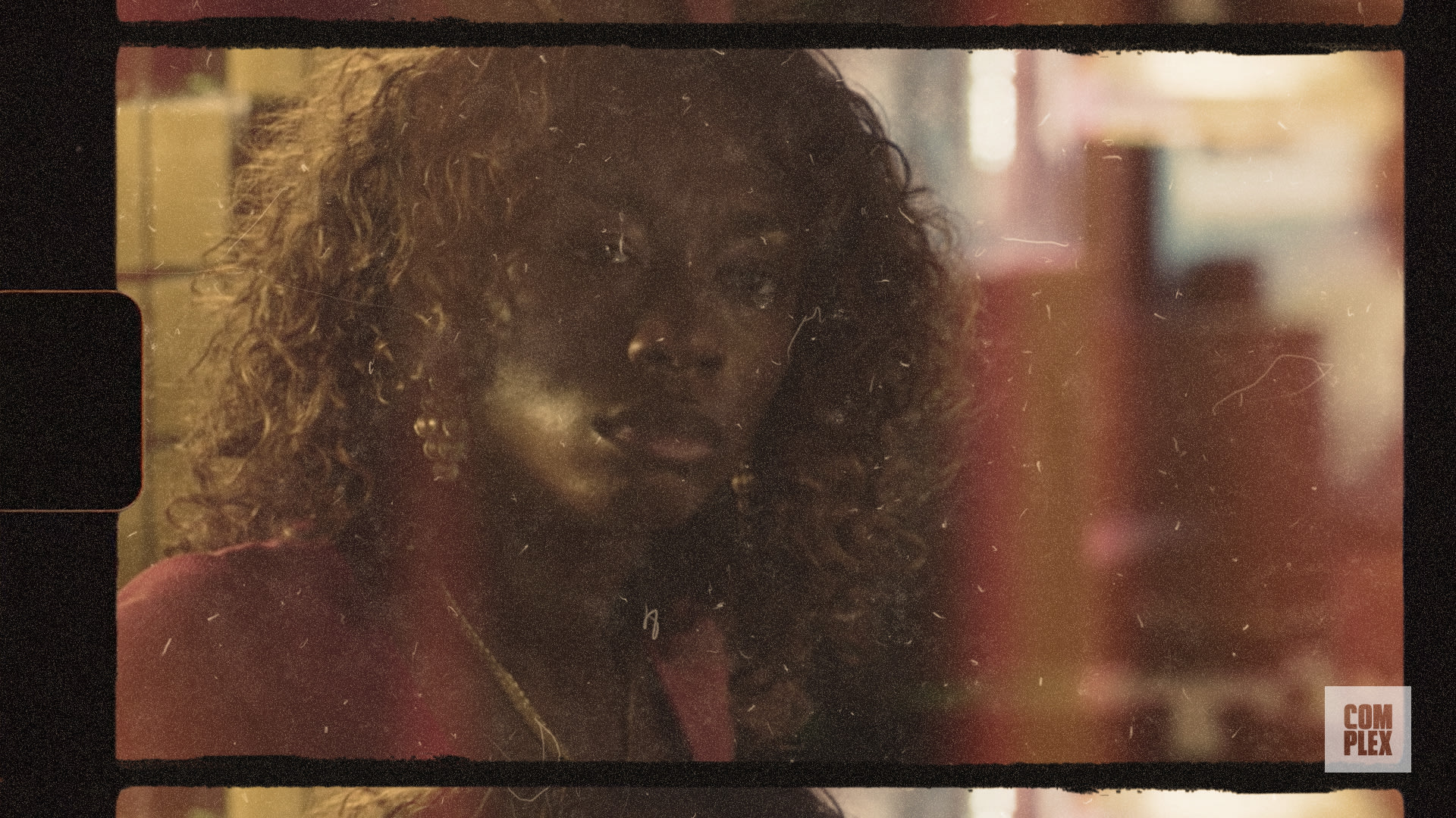 A person with curly hair is looking towards the camera in a dimly lit setting. The image has a vintage film effect.