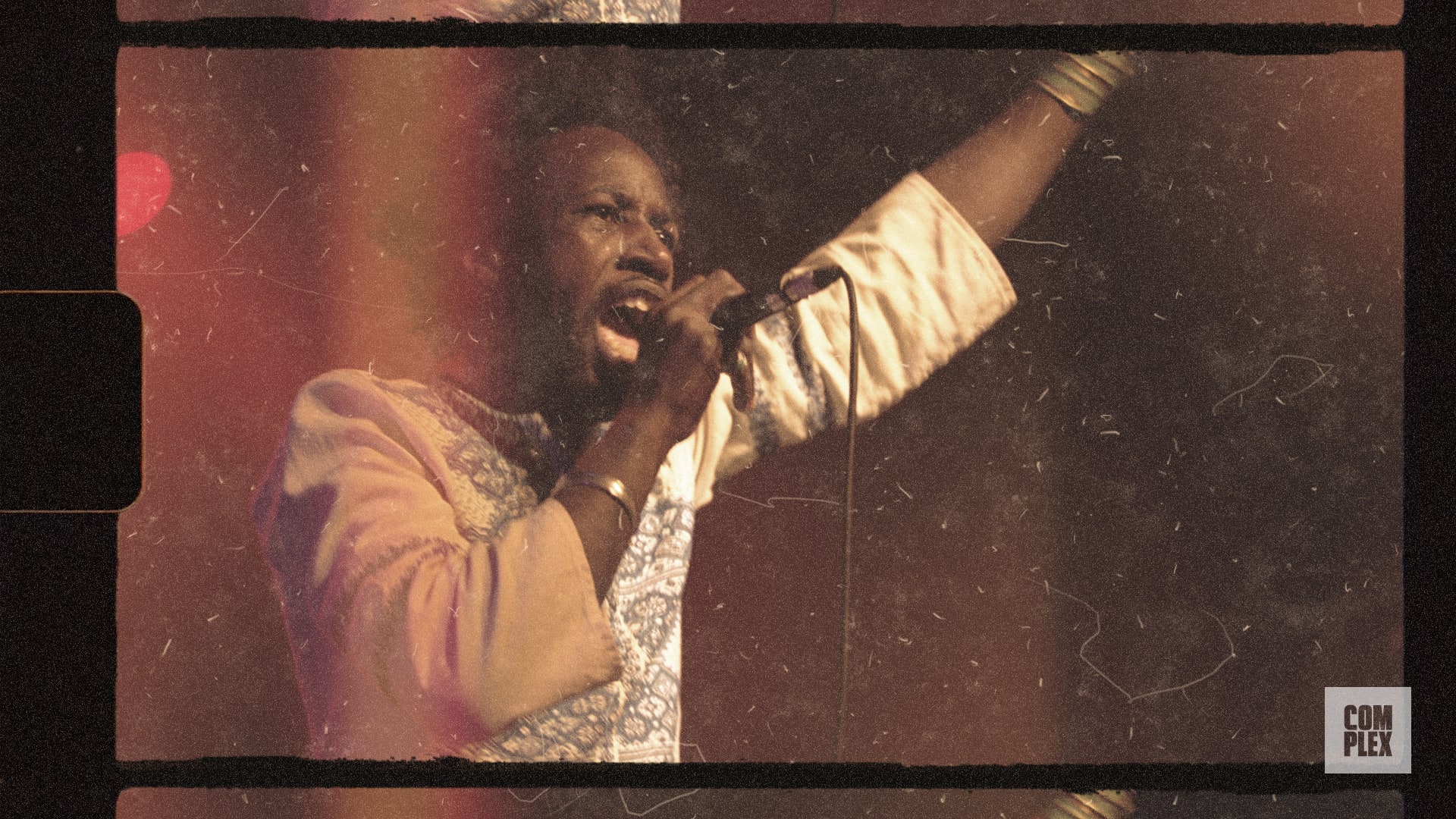 A performer passionately singing into a microphone on stage, with one arm raised. The image has a vintage, grainy effect.