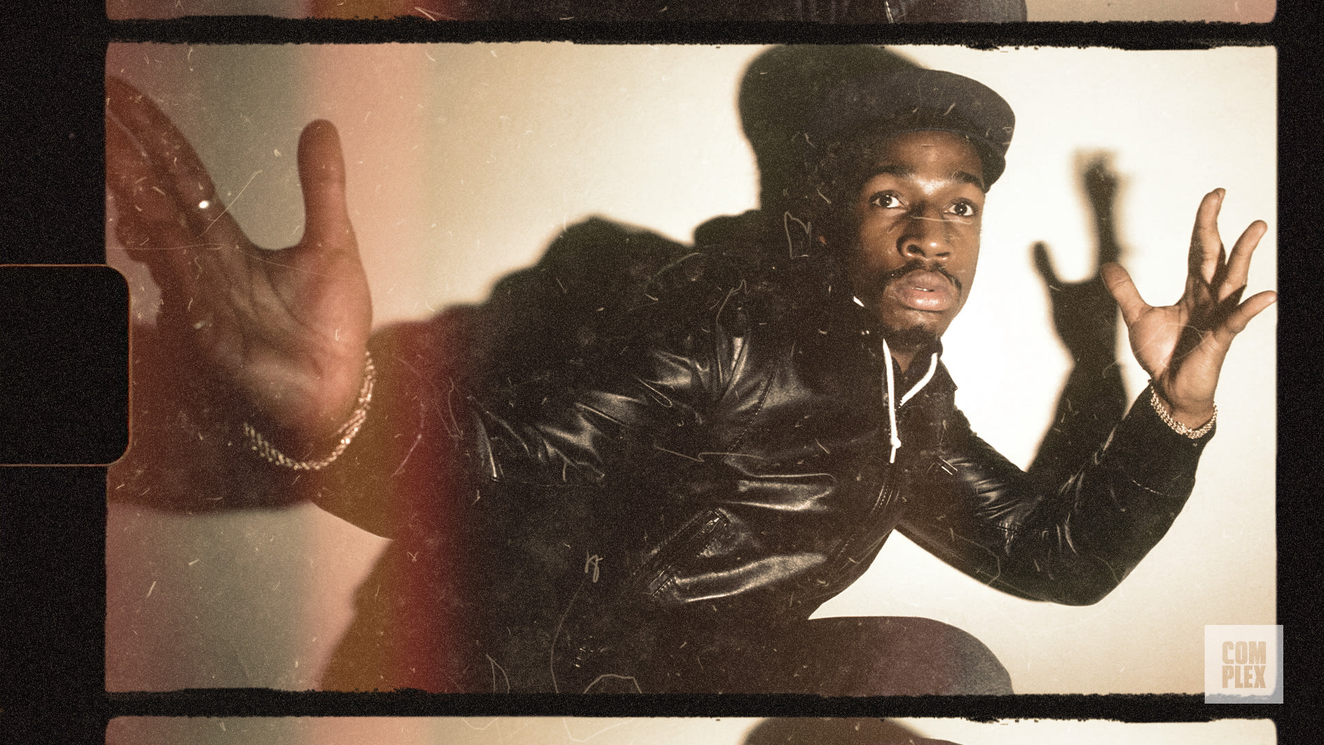 A man in a black leather jacket and cap poses dynamically against a light background with a film grain effect.
