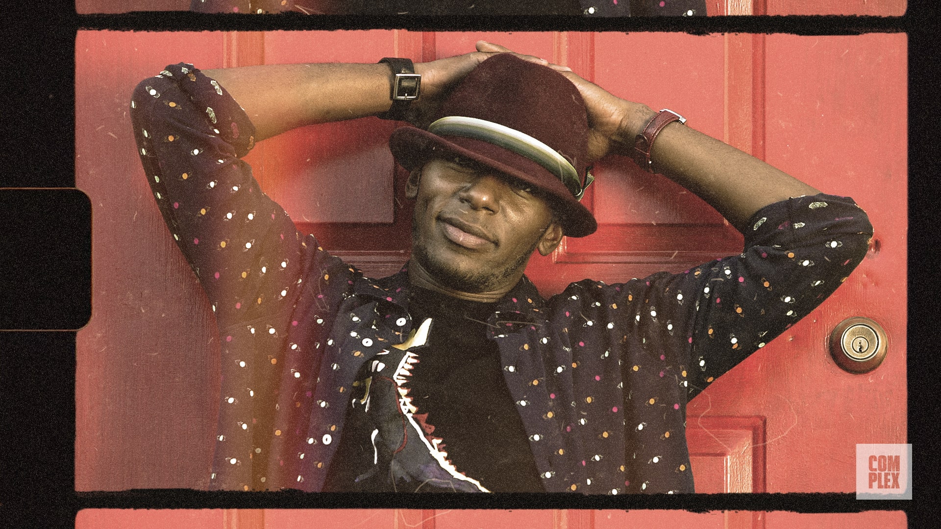 Man in a polka dot shirt and hat leaning against a red door, arms behind head, with a relaxed expression.