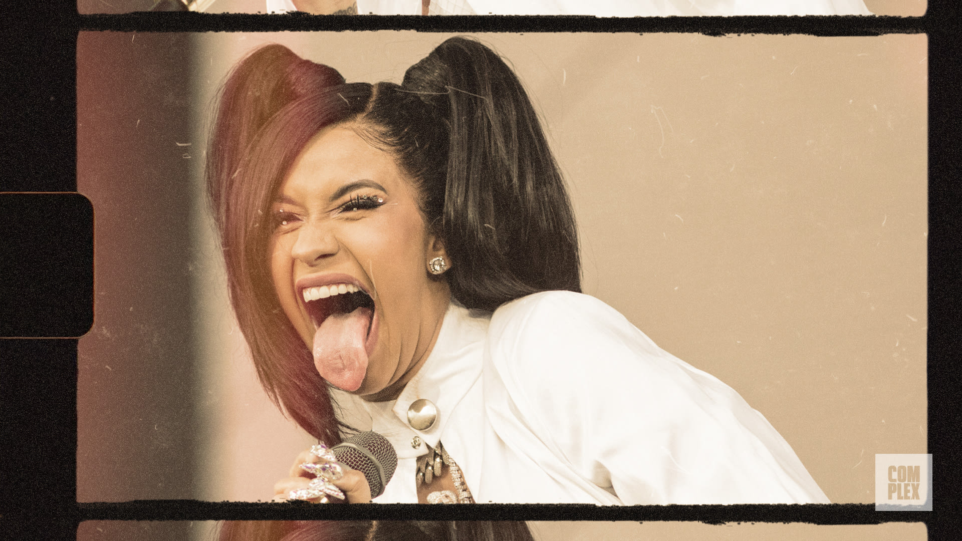 Cardi B playfully sticks out her tongue, holding a microphone, wearing a white outfit with pigtails.