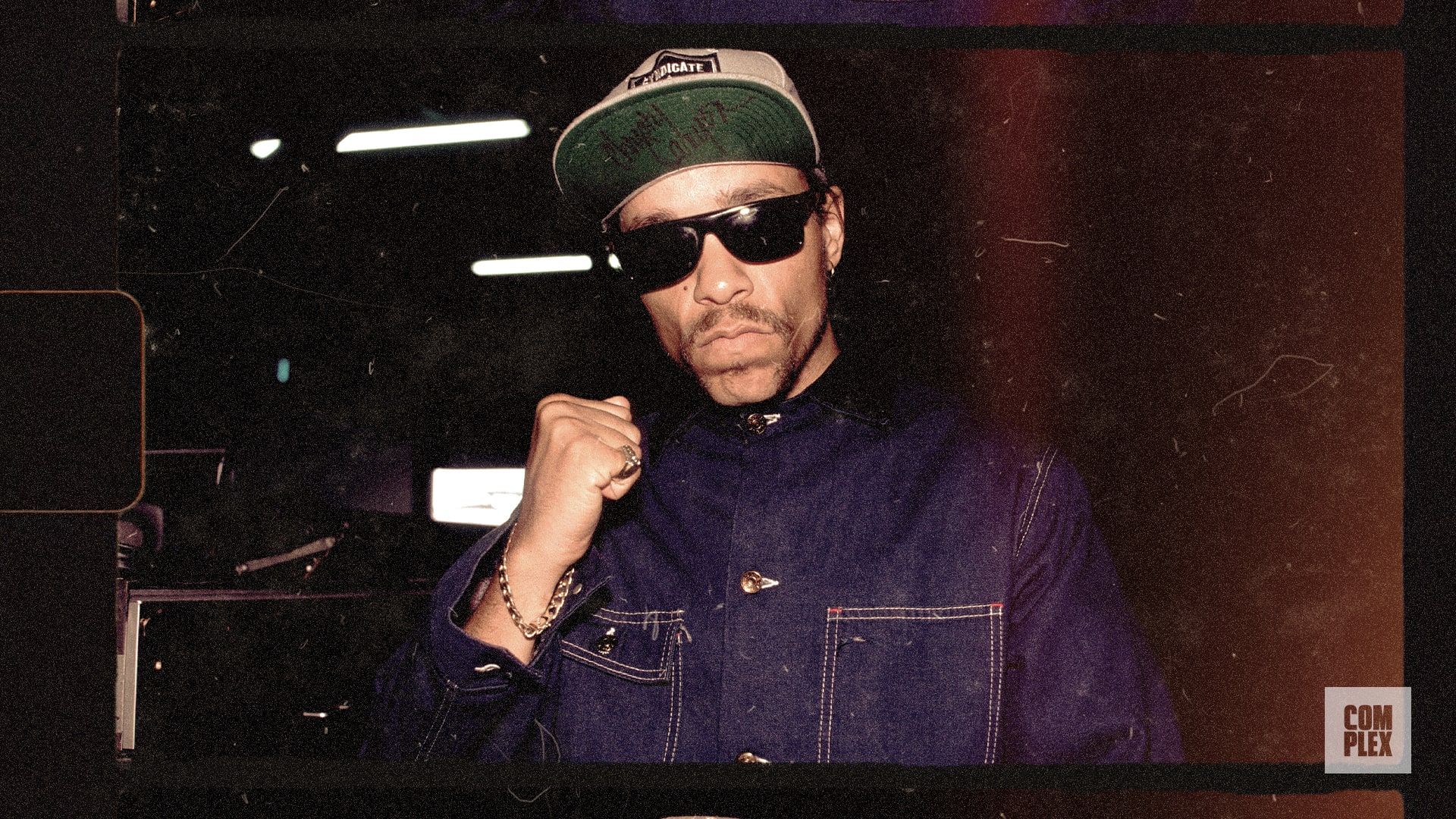 A person wearing sunglasses, a denim jacket, and a cap, posing with a fist raised. The image has a vintage filter.