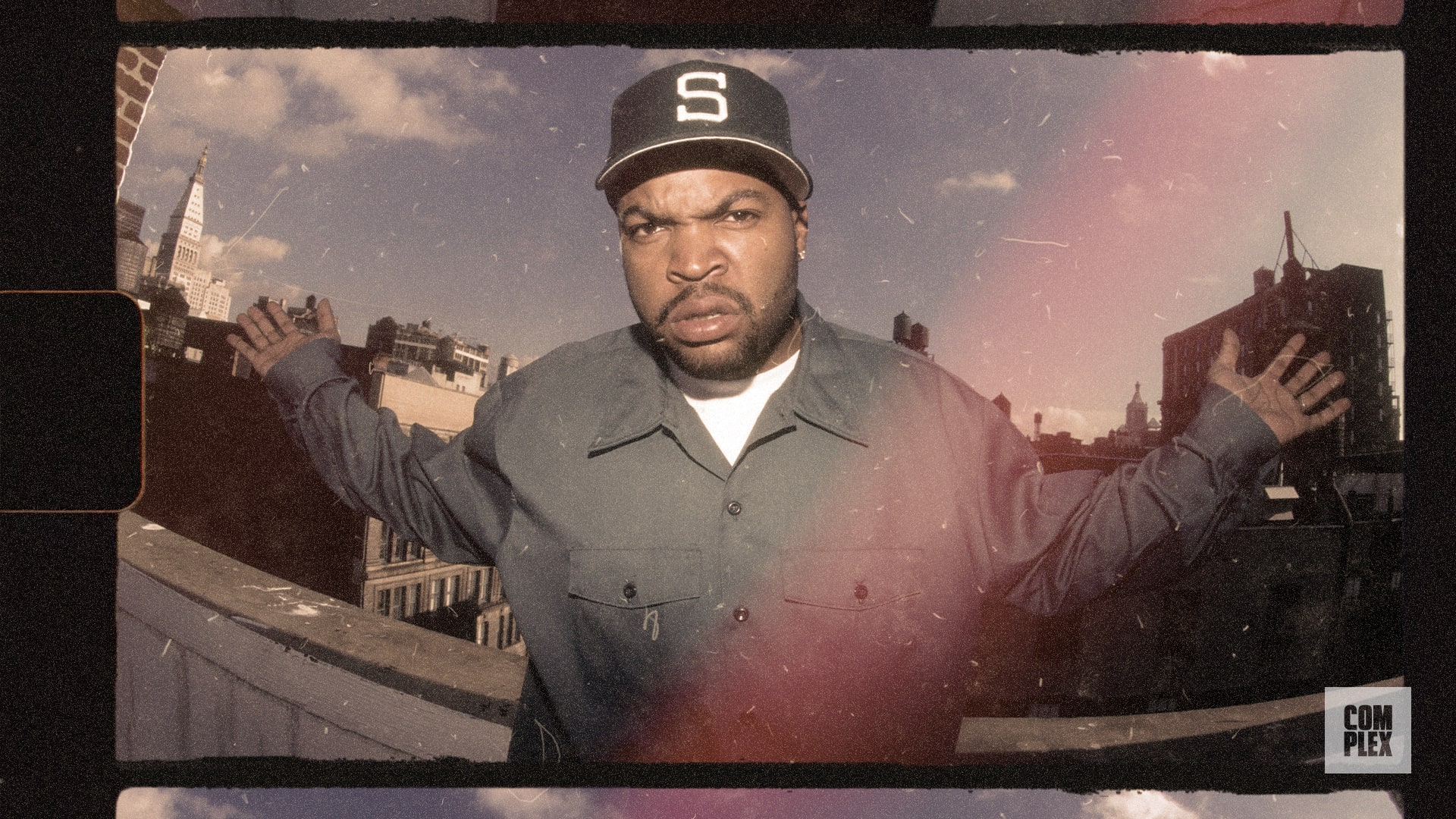 A man wearing a cap and jacket stands on a rooftop with a city skyline in the background, arms outstretched.